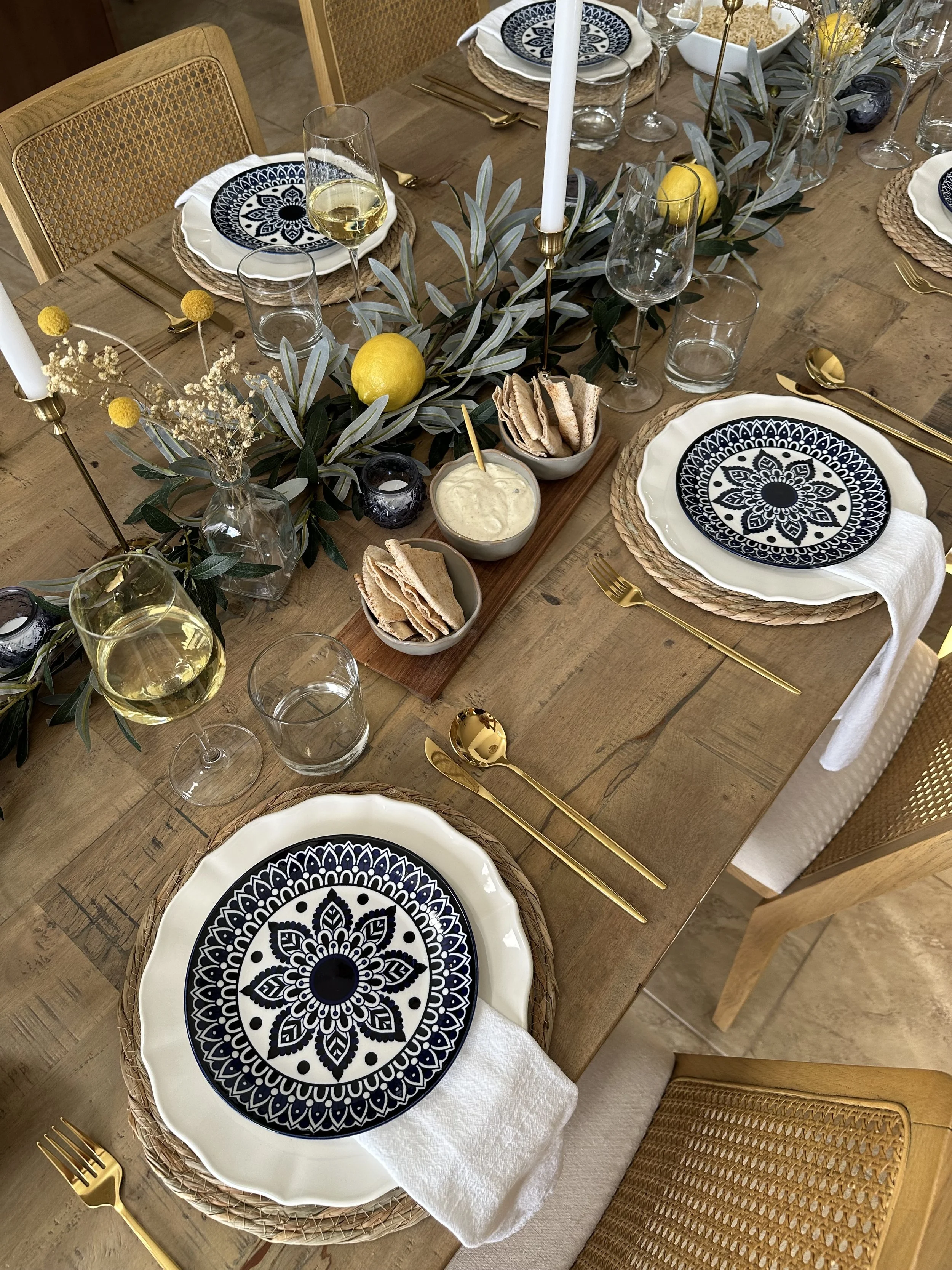 A Mediterranean-inspired dining tablescape rental for a summer dinner party. Includes olive leaf garland, lemons, blue and white floral plates, gold cutlery, candlesticks, and white linen napkins.