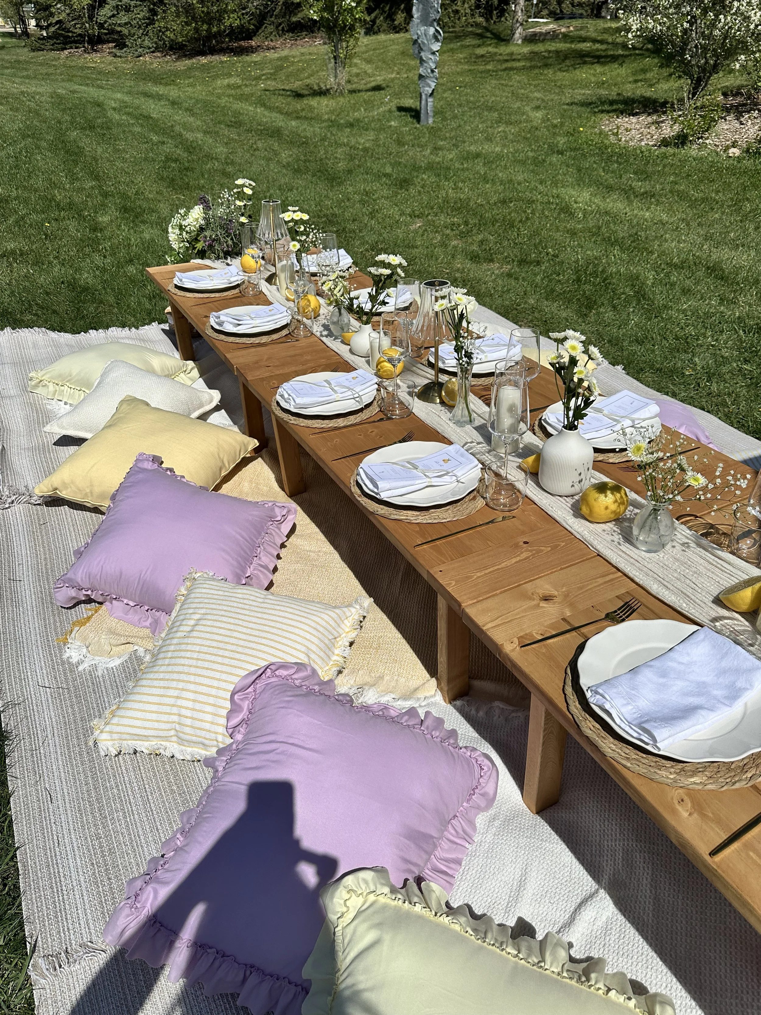 An outdoor dining setup on a wooden table decorated with flowers, candles, and lemons, placed on a large textured rug on the grass with pastel-colored pillows surrounding it.