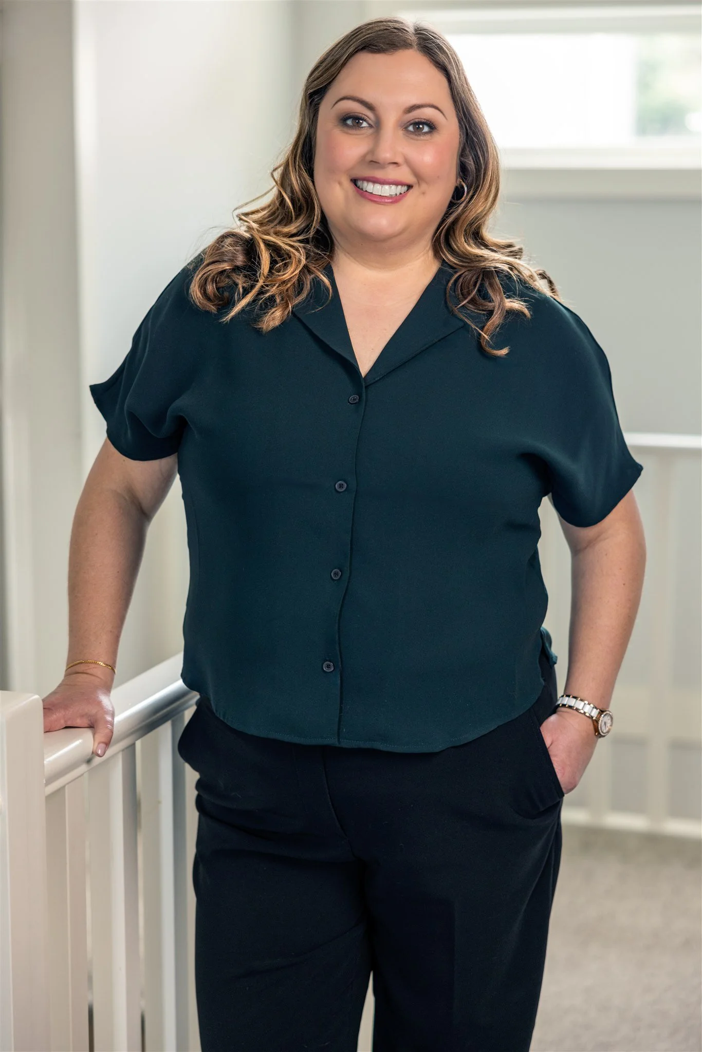 Shannon Shaughnessy, a woman with shoulder-length curly hair, wearing a black short-sleeve button-up shirt and black pants, standing indoors with her hand resting on a white railing, smiling at the camera.