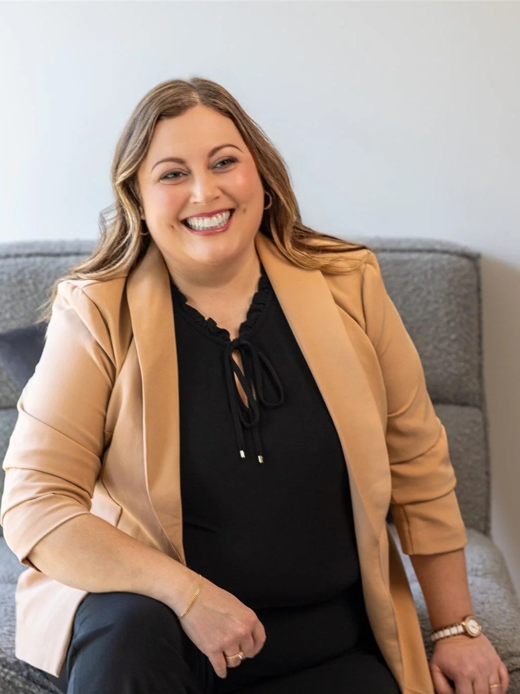 Shannon Shaughnessy, a smiling woman with shoulder-length brown hair, wearing a black blouse and a beige blazer, sitting on a gray couch in a neutral room.