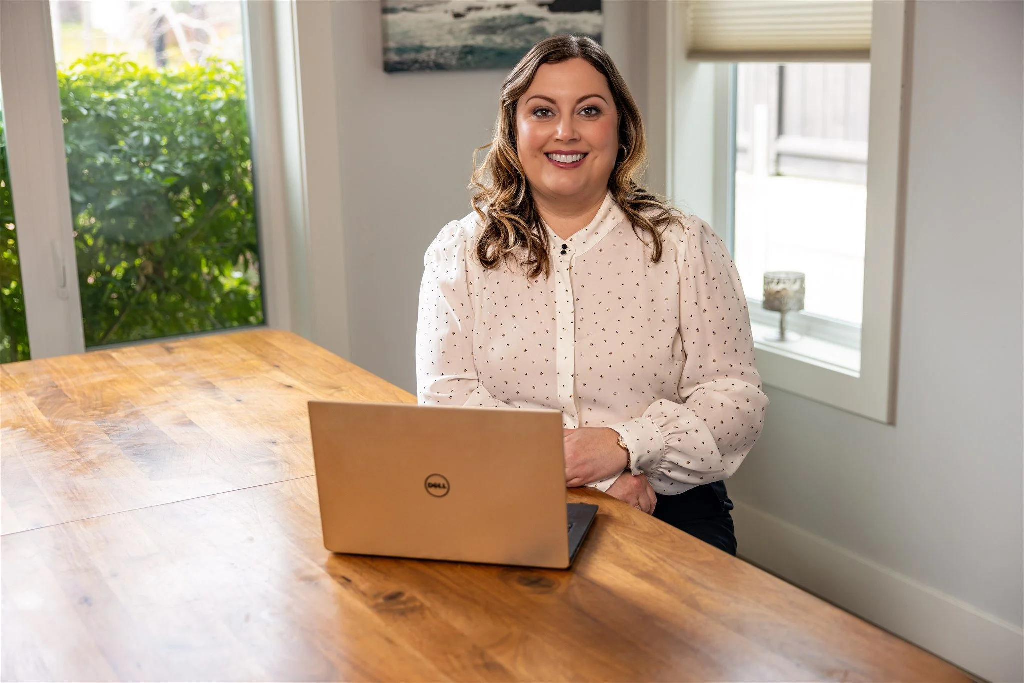 A woman with brown, wavy hair in a white blouse with small black dots, sitting at a wooden table with a Dell laptop in front of her, in a room with white walls, two windows, and a painting hanging on the wall.