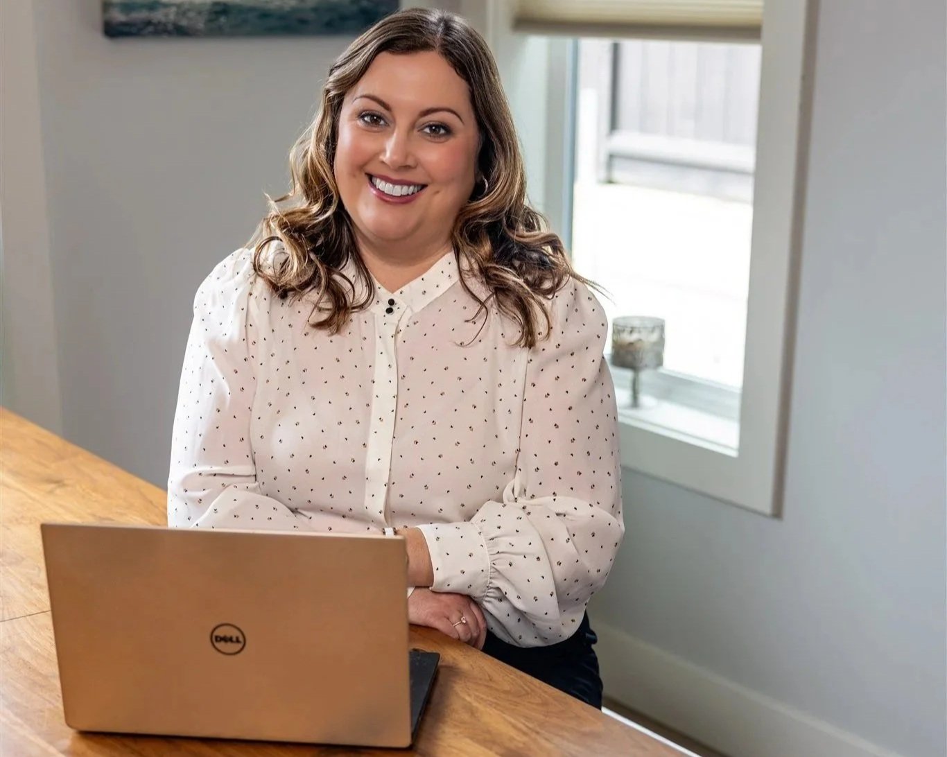 Shannon Shaughnessy, a woman with wavy brown hair smiling at the camera, sitting at a wooden table with a gold Dell laptop in front of her in a bright room with a window in the background.