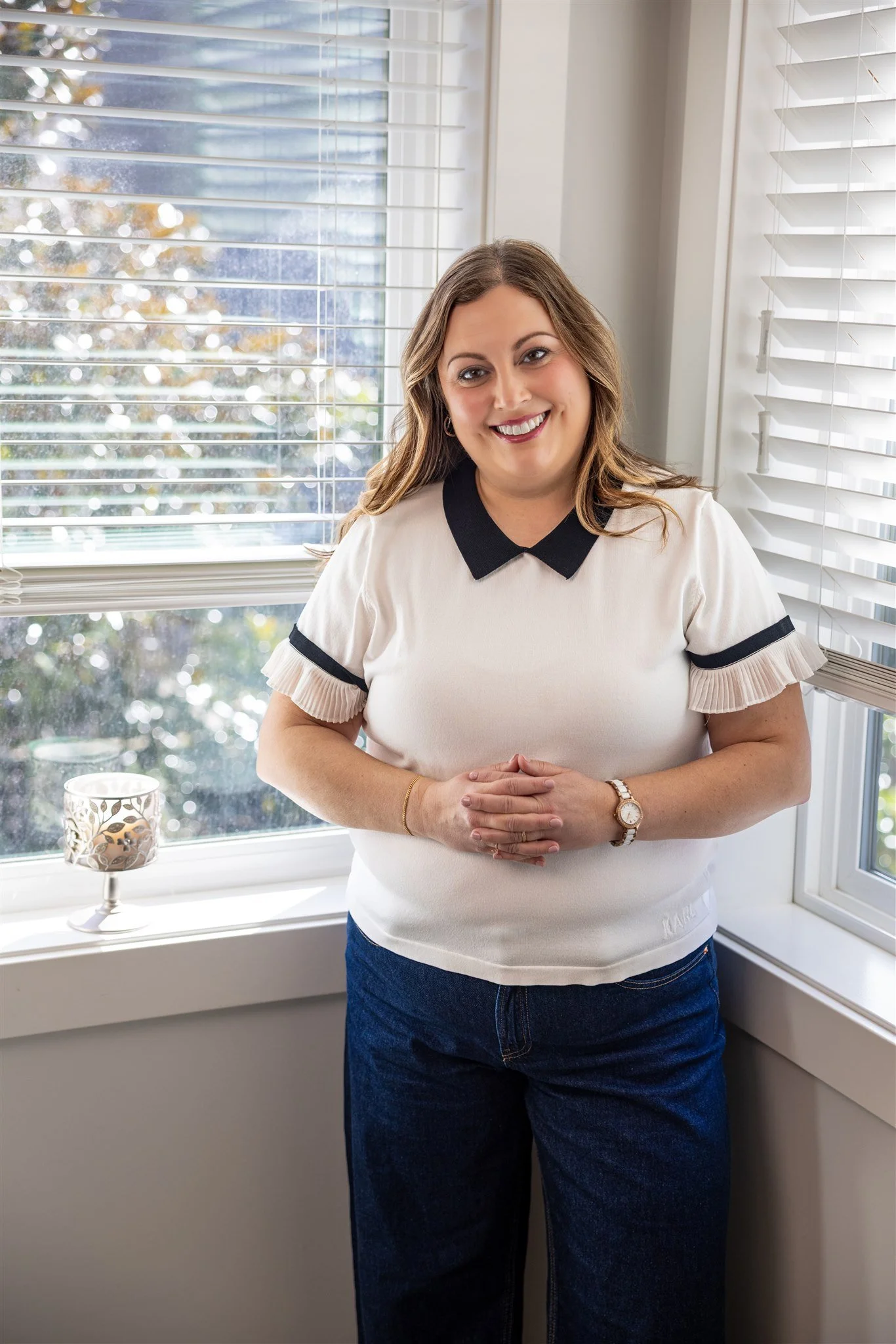 Shannon Shaughnessy, a woman with long, wavy hair, smiling, stands indoors near a window with white blinds, wearing a white short-sleeved shirt with black accents and dark jeans.