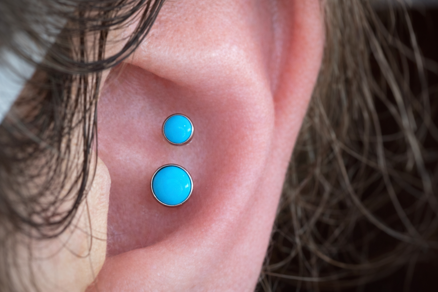Close-up of a person's face showing curly brown hair and two blue circular facial piercings on the cheek.