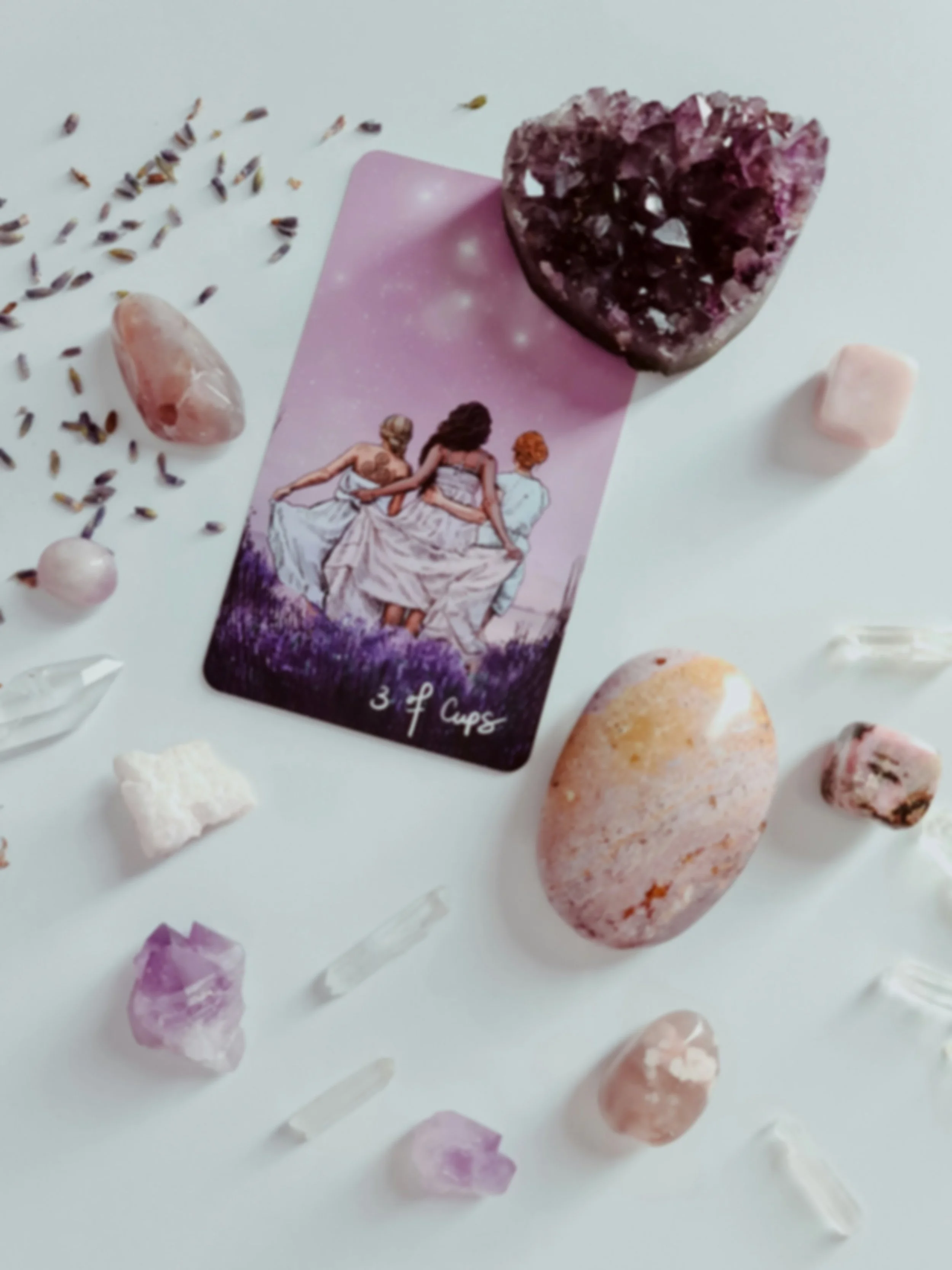 A tarot card labeled "3 of Cups" surrounded by various gemstones including amethysts, rose quartz, and other crystals on a white surface.