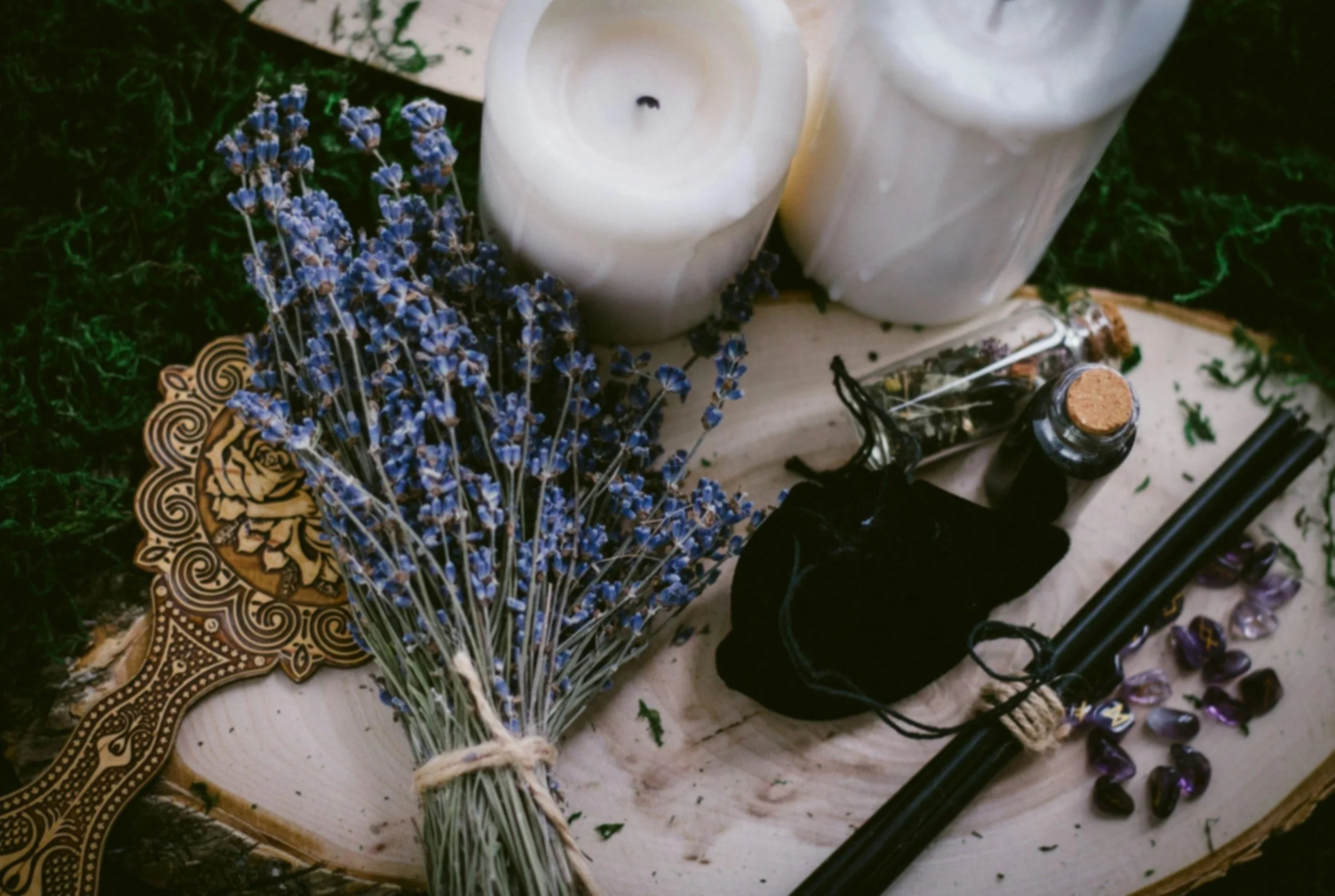Arrangement of lavender flowers, white candles, small glass vial, black cloth pouch, tarot card, black pen, and purple stones on a wooden surface with green moss background.