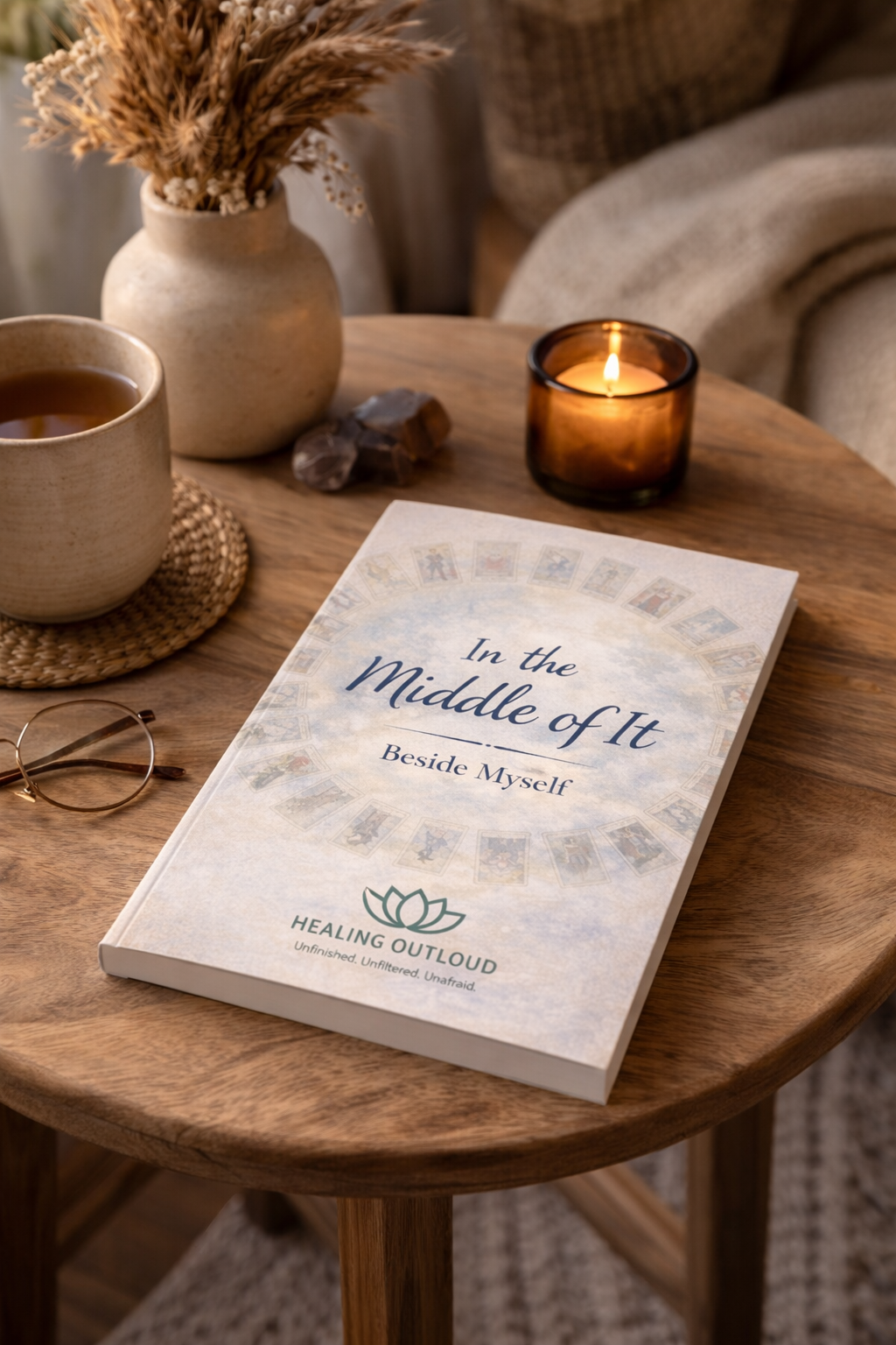 A wooden table with a book titled 'In the Middle of It Beside Myself' by Healing Outloud, a cup of tea on a woven coaster, a candle in a brown glass holder, a vase with dried wheat, and a pair of glasses.