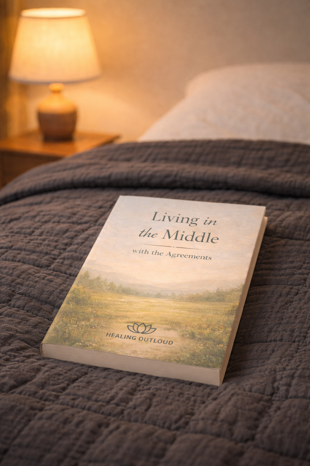 A paperback book titled 'Living in the Middle with the Agreements' resting on a dark gray quilted bedspread in a bedroom with a lit table lamp in the background.