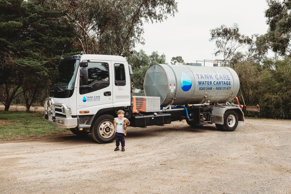 A young boy standing in front of a water delivery truck labeled 'Tank Care Water Cartage' parked on a dirt area with trees in the background. Adelaide Water supplier water truck.