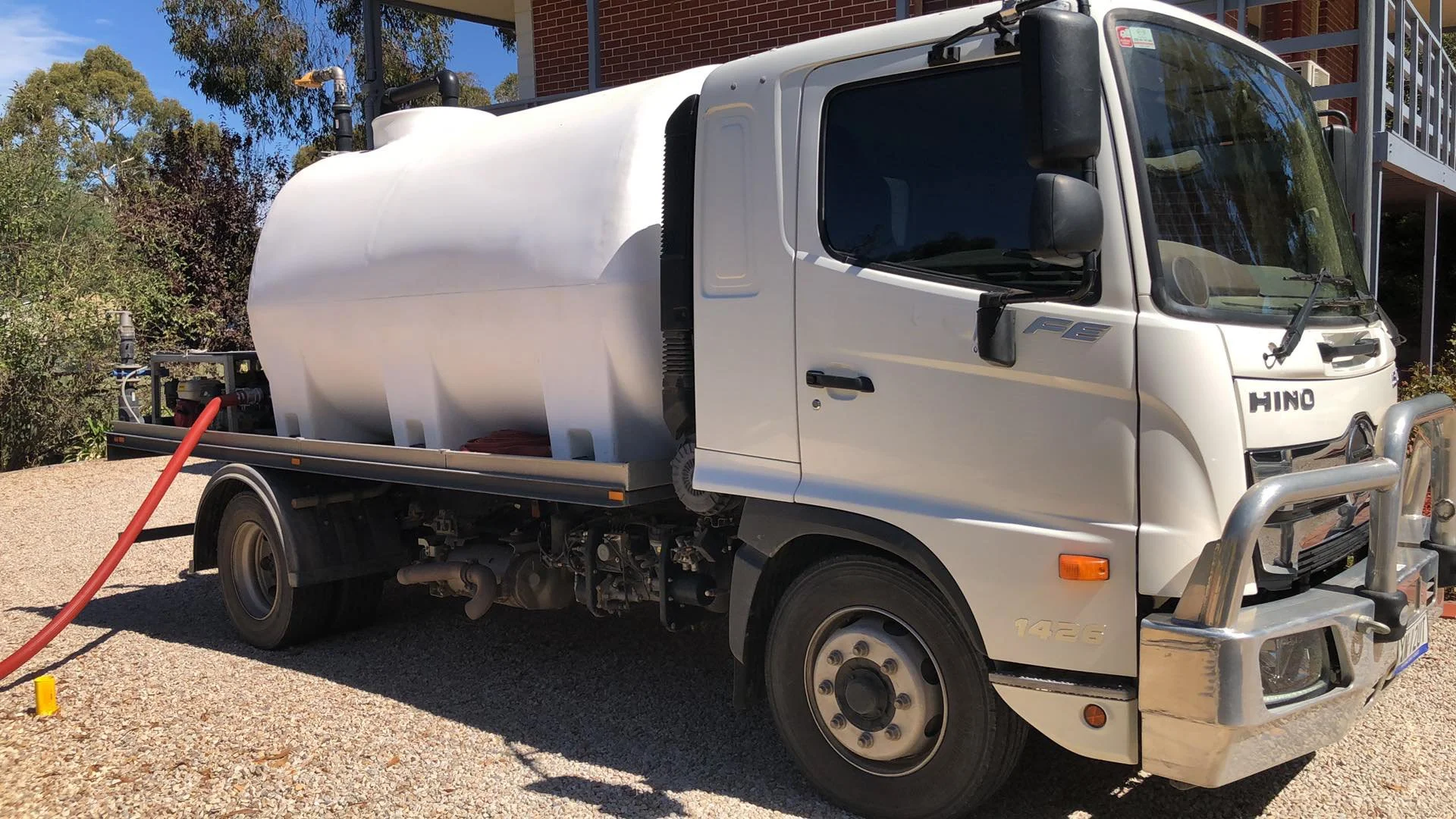 White water tanker truck parked on gravel with a red hose connected to it, delivering water to a domestic customer. greenery and a brick building in the background.