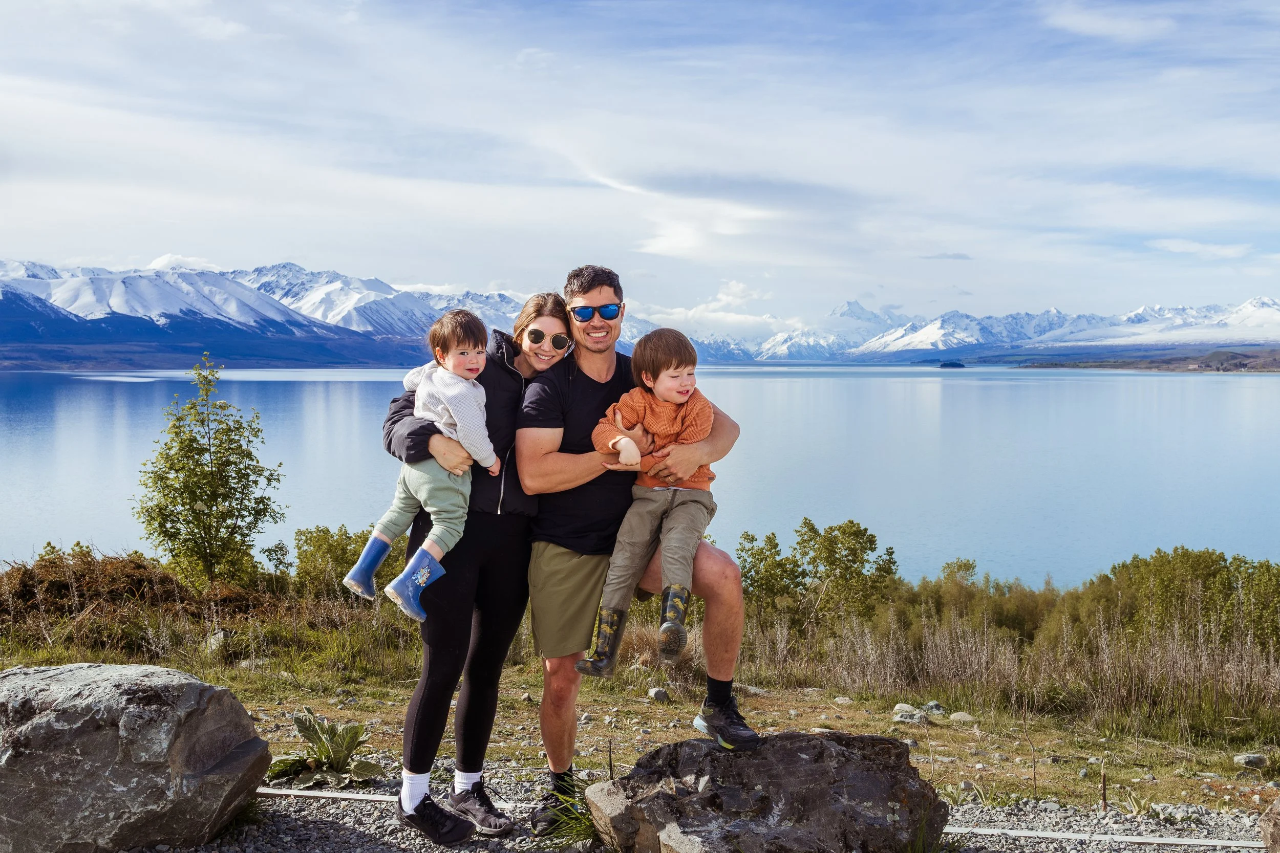 Family of four enjoying outdoors by a lake with snow-capped mountains in the background. The father and mother are smiling, with the mother holding a young child and the father lifting another child. They are dressed in casual outdoor clothing, with the children wearing rain boots.