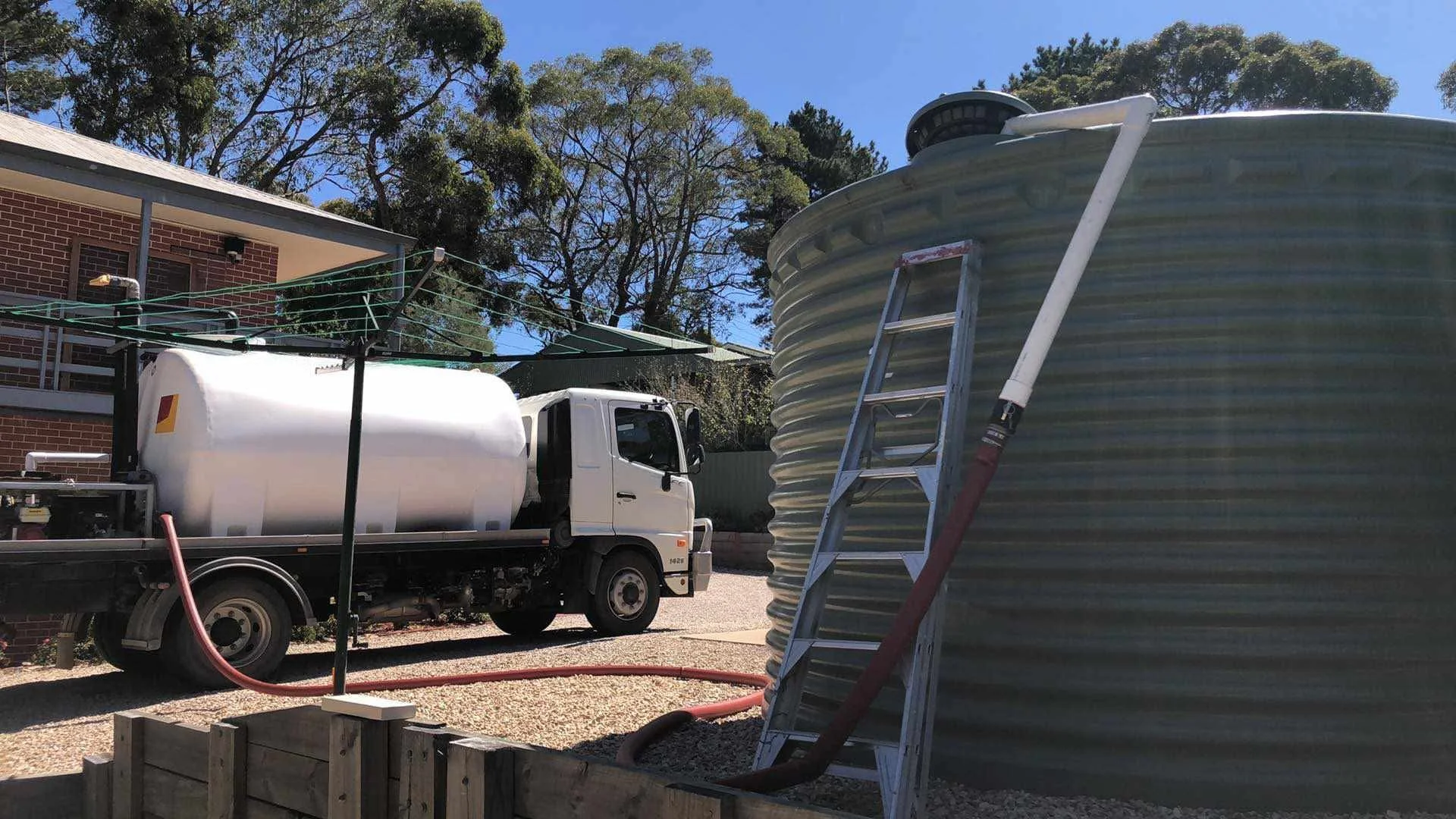 A white water truck with a tank, a ladder, and a large cylindrical gray water tank next to a house, with trees and blue sky in the background.
