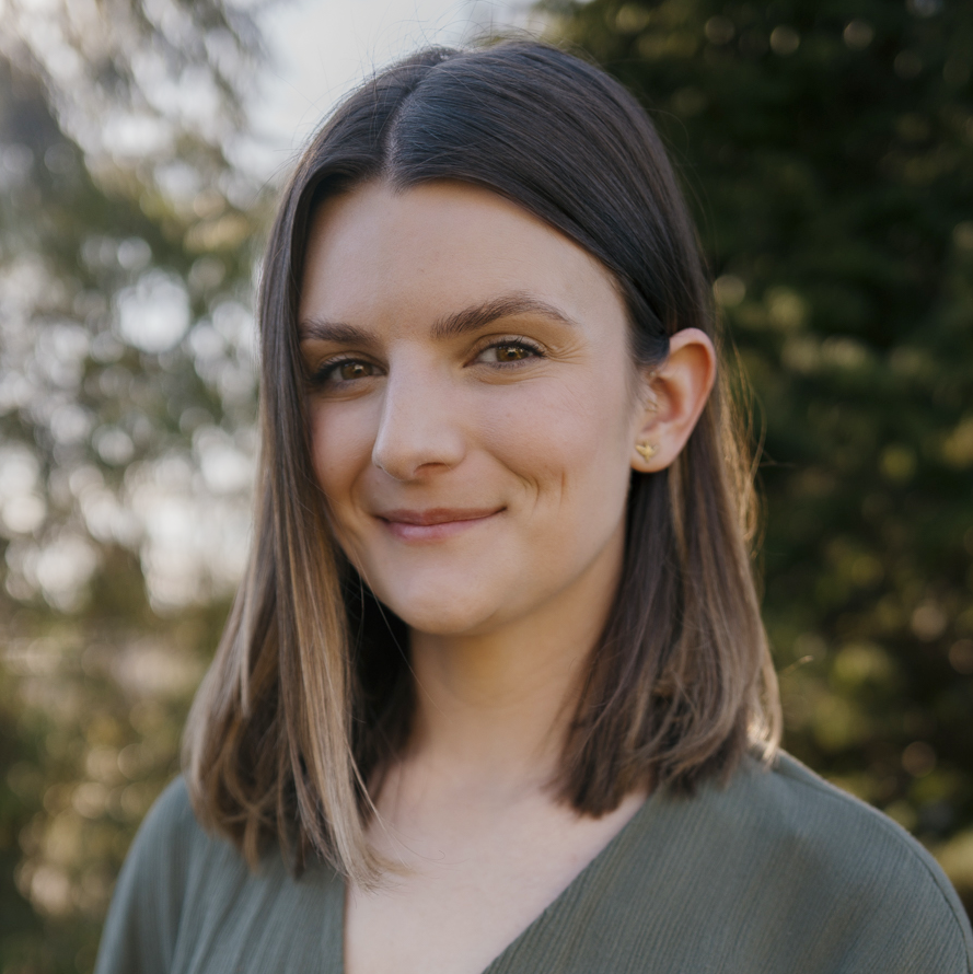 A woman with shoulder-length brown hair smiling outdoors with blurred trees in the background.