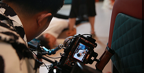 Person operating a camera filming a scene with a person lying on the floor in an indoor setting.