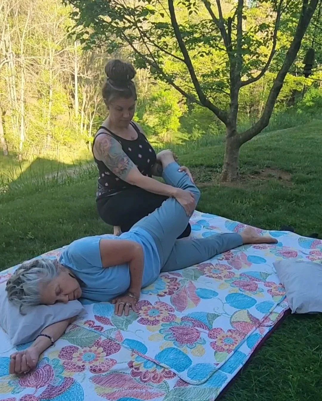 A woman with tattoos giving a Thai massage outdoors on a blanket  with trees and green grass all around them.