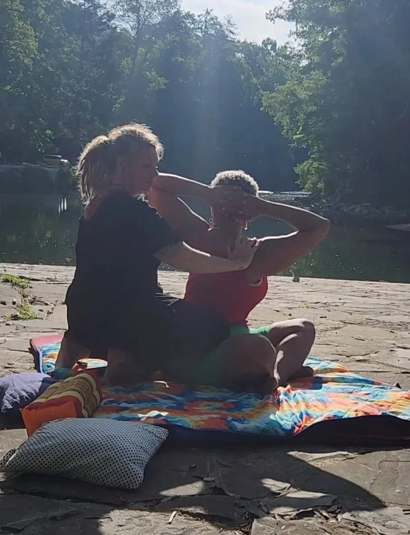 Two people are practicing Thai Massage on a colorful blanket by a river, with trees in the background and sunlight filtering through the trees.