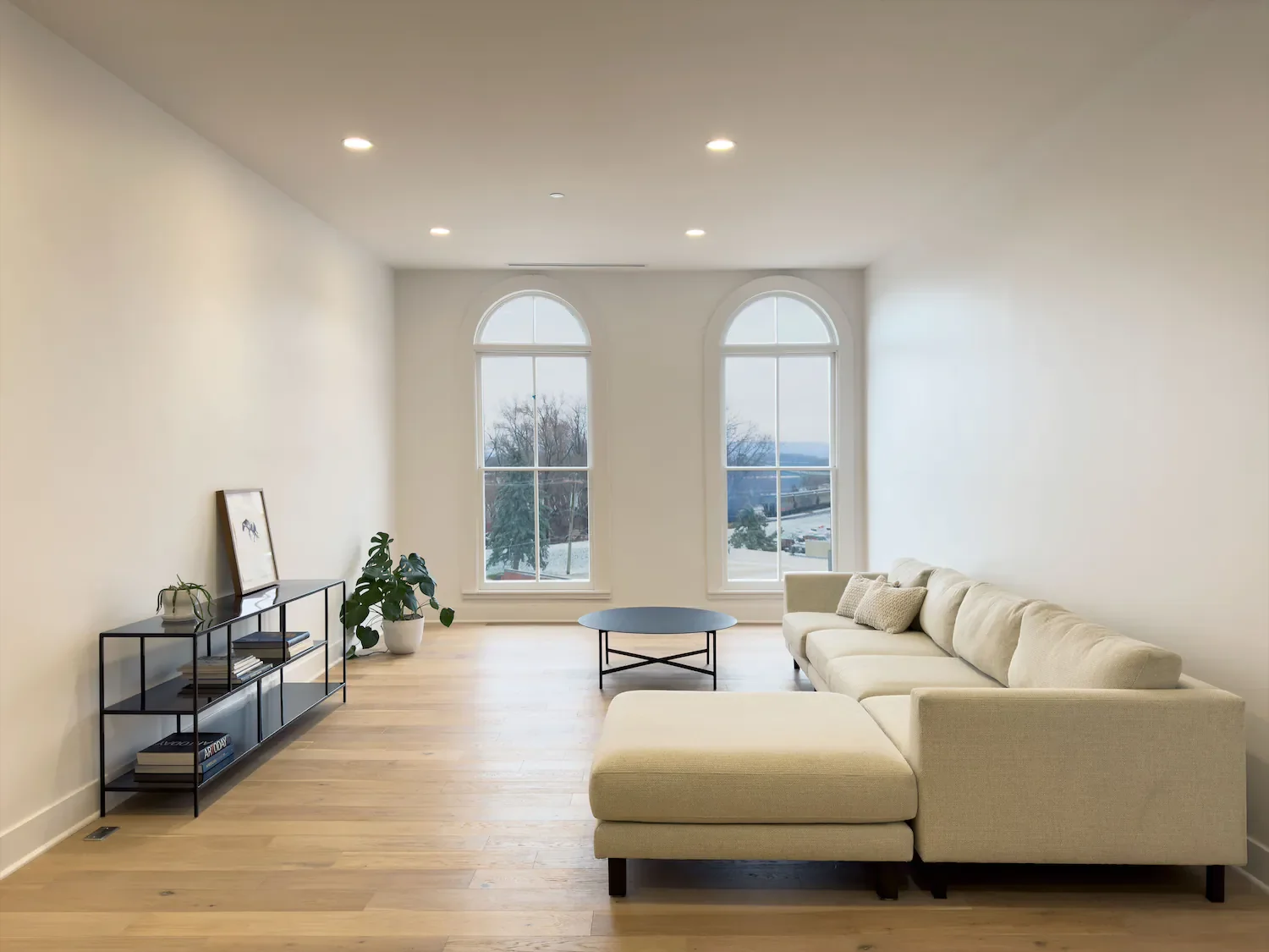 Minimalist living room with a large beige sectional sofa, black coffee table, black metal shelf with books, and a potted plant, illuminated by natural light from two tall arched windows.
