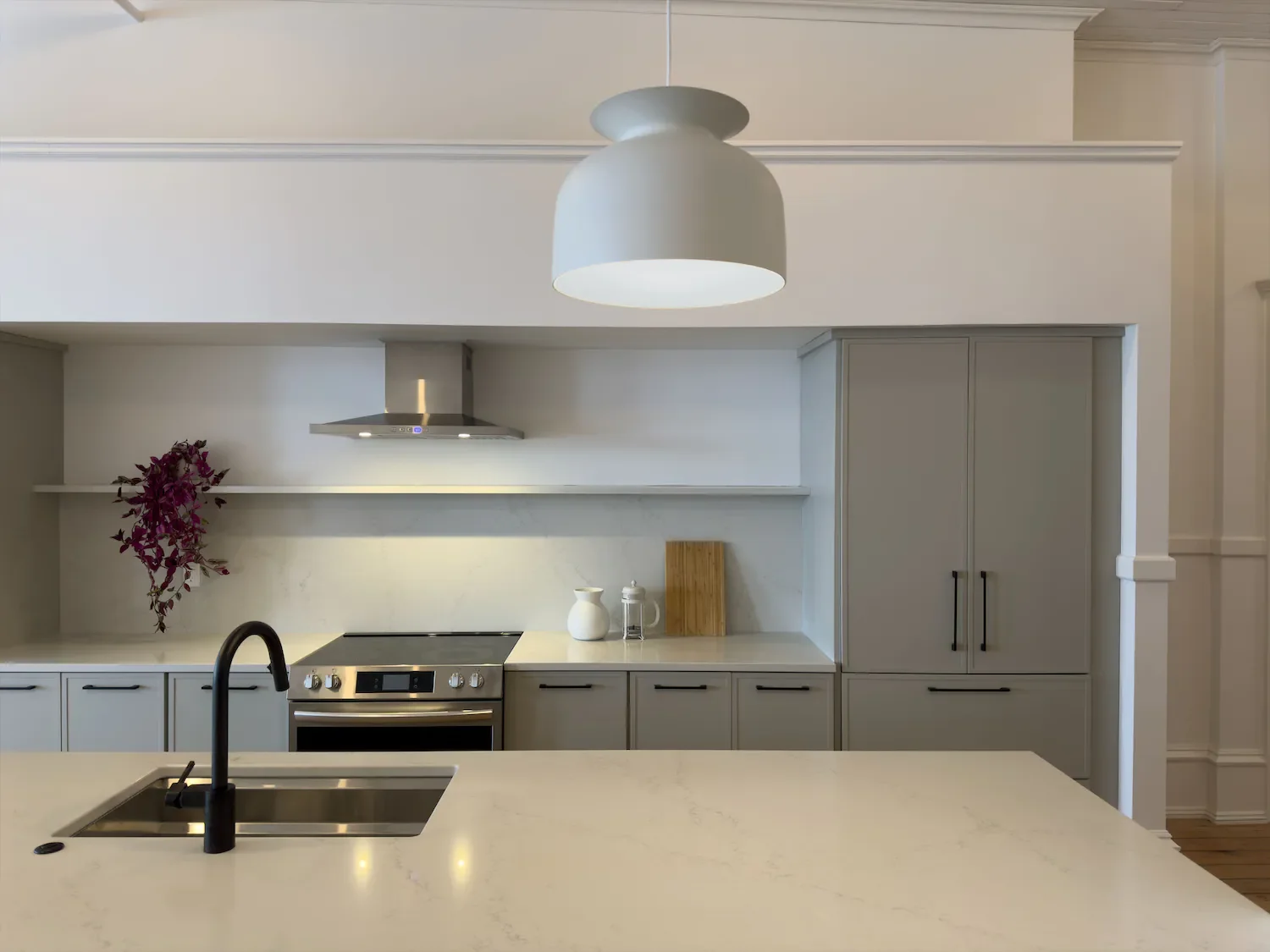 Modern kitchen with light gray cabinets, white countertops, a black faucet, a built-in black stovetop, stainless steel range hood, and a white vase on the counter. There is a white pendant light hanging from the ceiling.