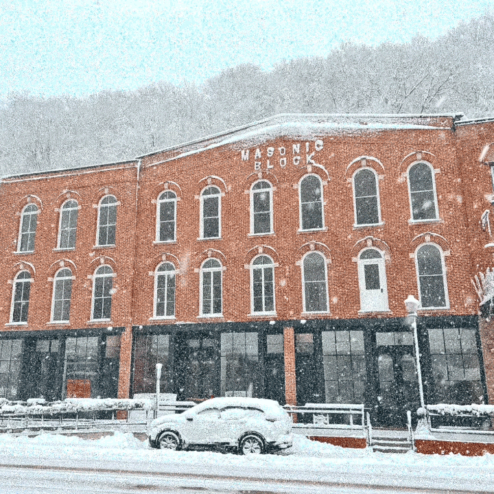 Snow-covered street with parked car in front of a red brick building labeled Masonic Block, snow falling, winter scene