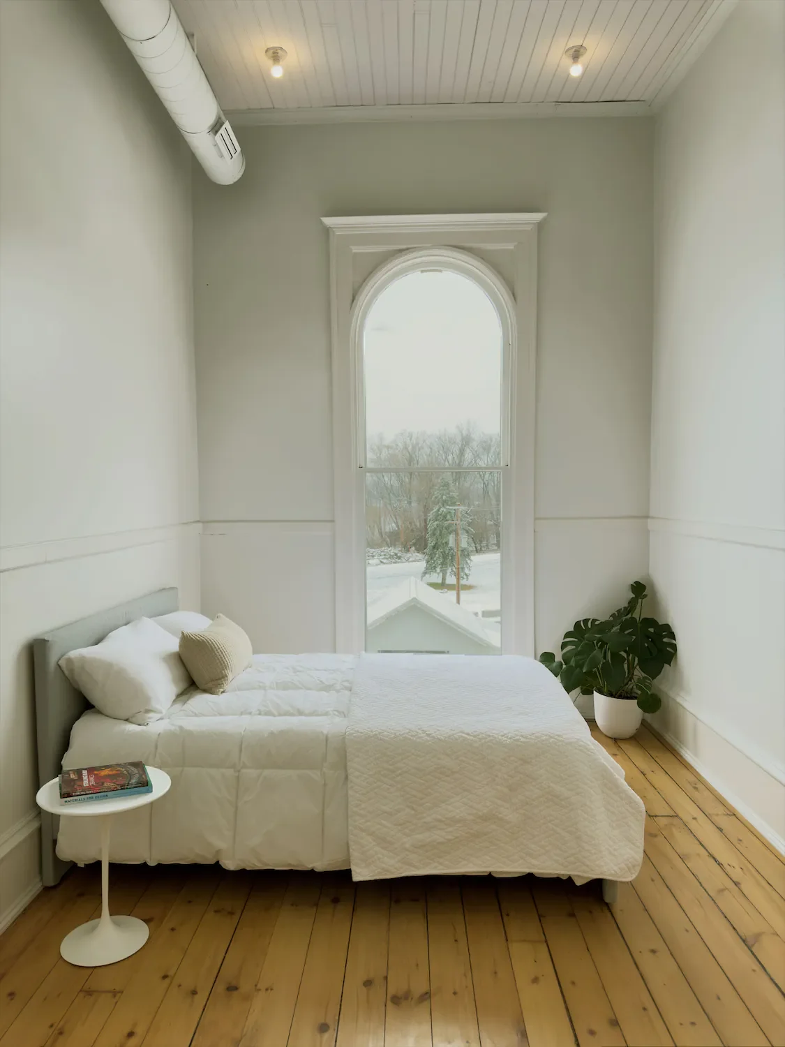 Minimalist bedroom with white bedding, a small side table with books, a large arched window with a view of snow and trees, wooden floor, and a potted plant in the corner.