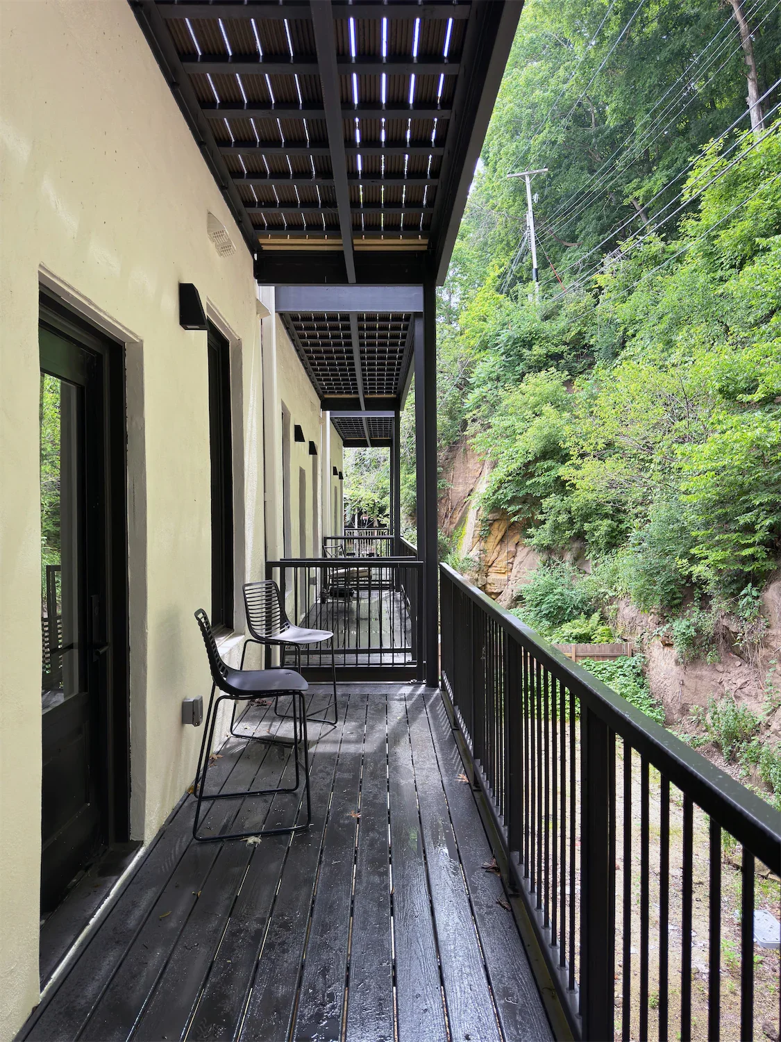 A corridor on a balcony with black chairs and a black railing outside a building, overlooking a green hillside with trees and power lines.