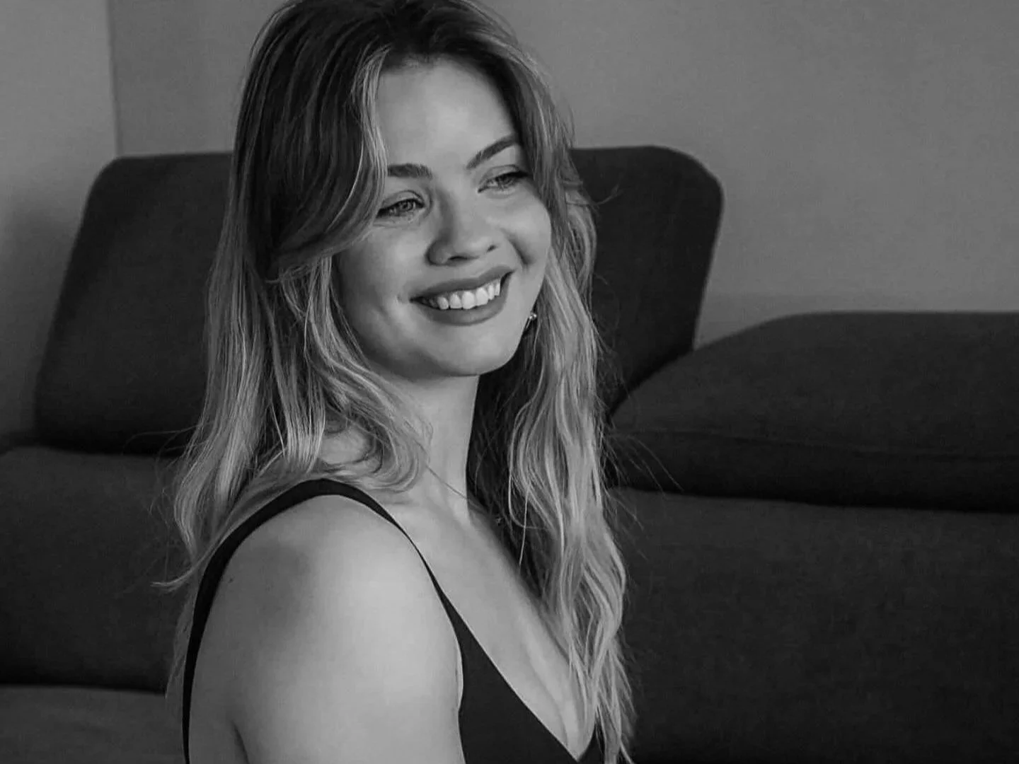 A smiling woman with long wavy hair and light skin, wearing a sleeveless top, sitting on a couch in a black-and-white photo.
