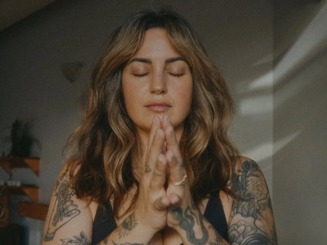 A woman with brown, wavy hair and tattoos on her arms and shoulders has eyes closed, hands pressed together in prayer or meditation, in a calm, indoor setting.