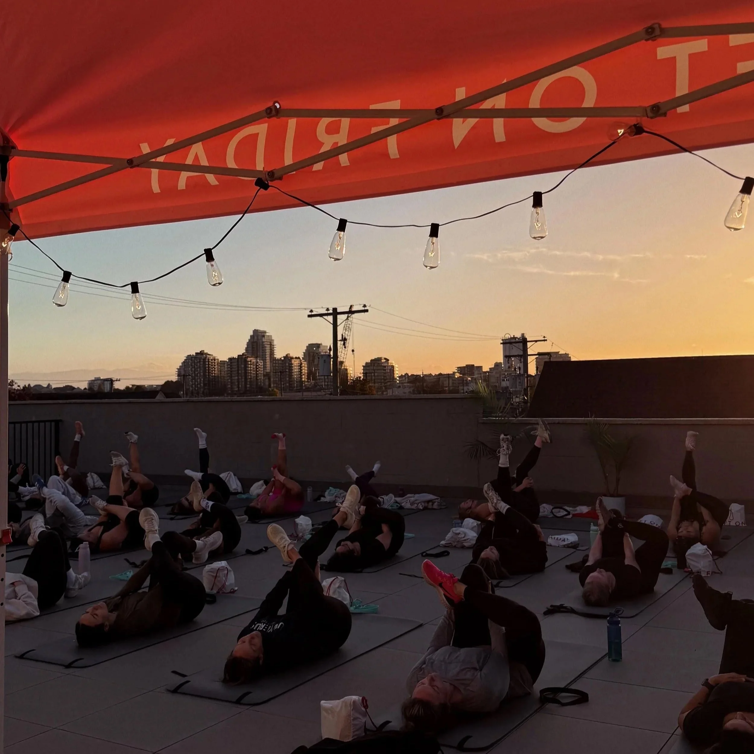 Group of people practicing yoga on a rooftop during sunset with city skyline in the background, illuminated by string lights.