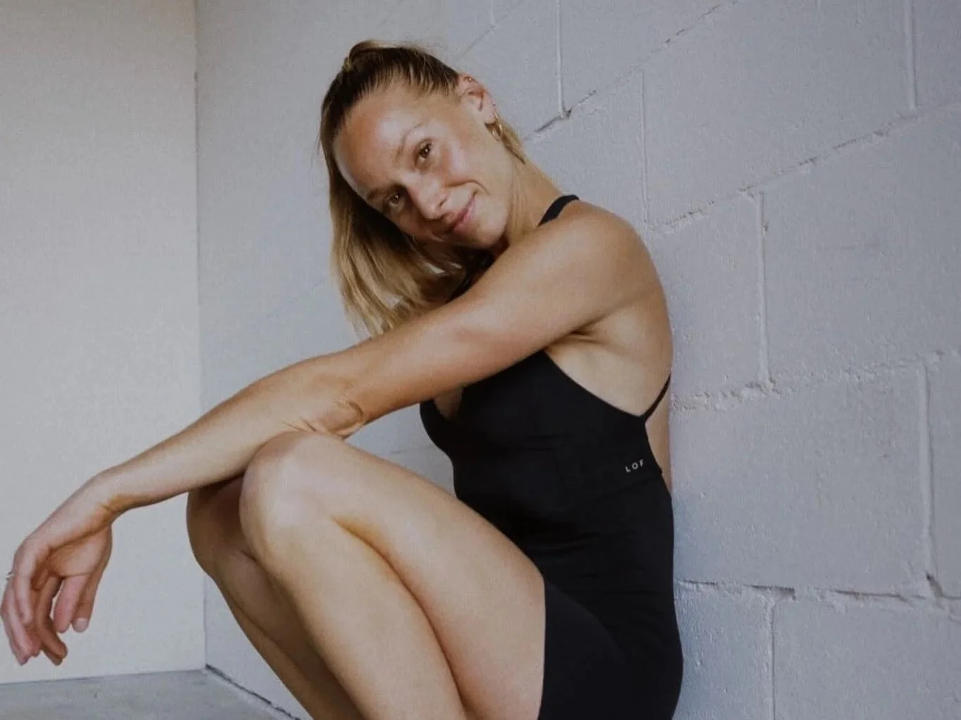 Woman sitting on the floor against a gray concrete wall, wearing a black tank top and shorts, smiling at the camera.