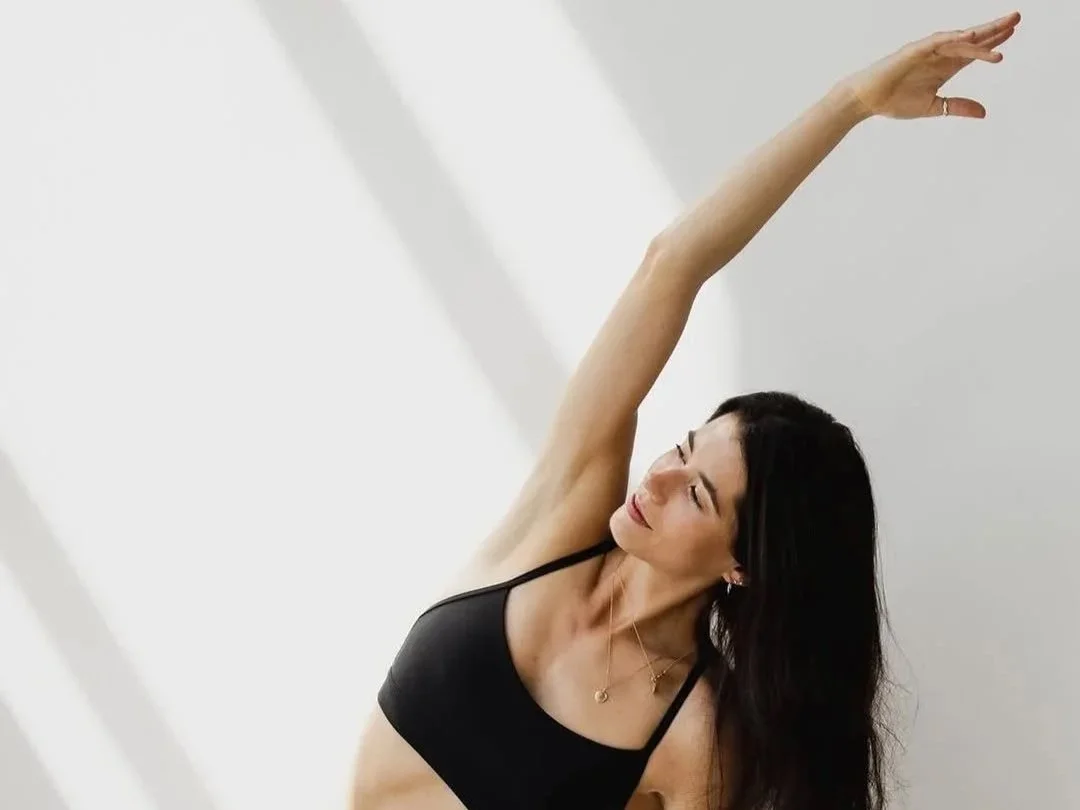 A woman with long dark hair practicing a side stretch yoga pose in a well-lit room, wearing a black sports bra.