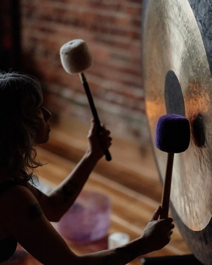A woman with curly hair striking a gong with a purple mallet, and a second woman with a puffed-up mallet hitting a large gong, both in a dimly lit room with exposed brick walls.
