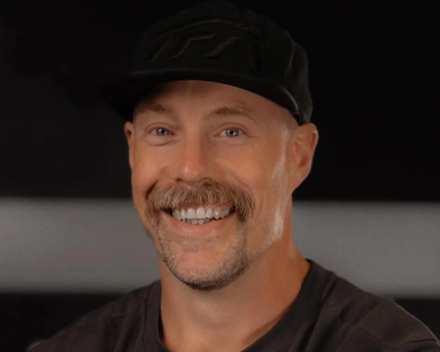 A man with a mustache and beard wearing a black cap smiling at the camera.