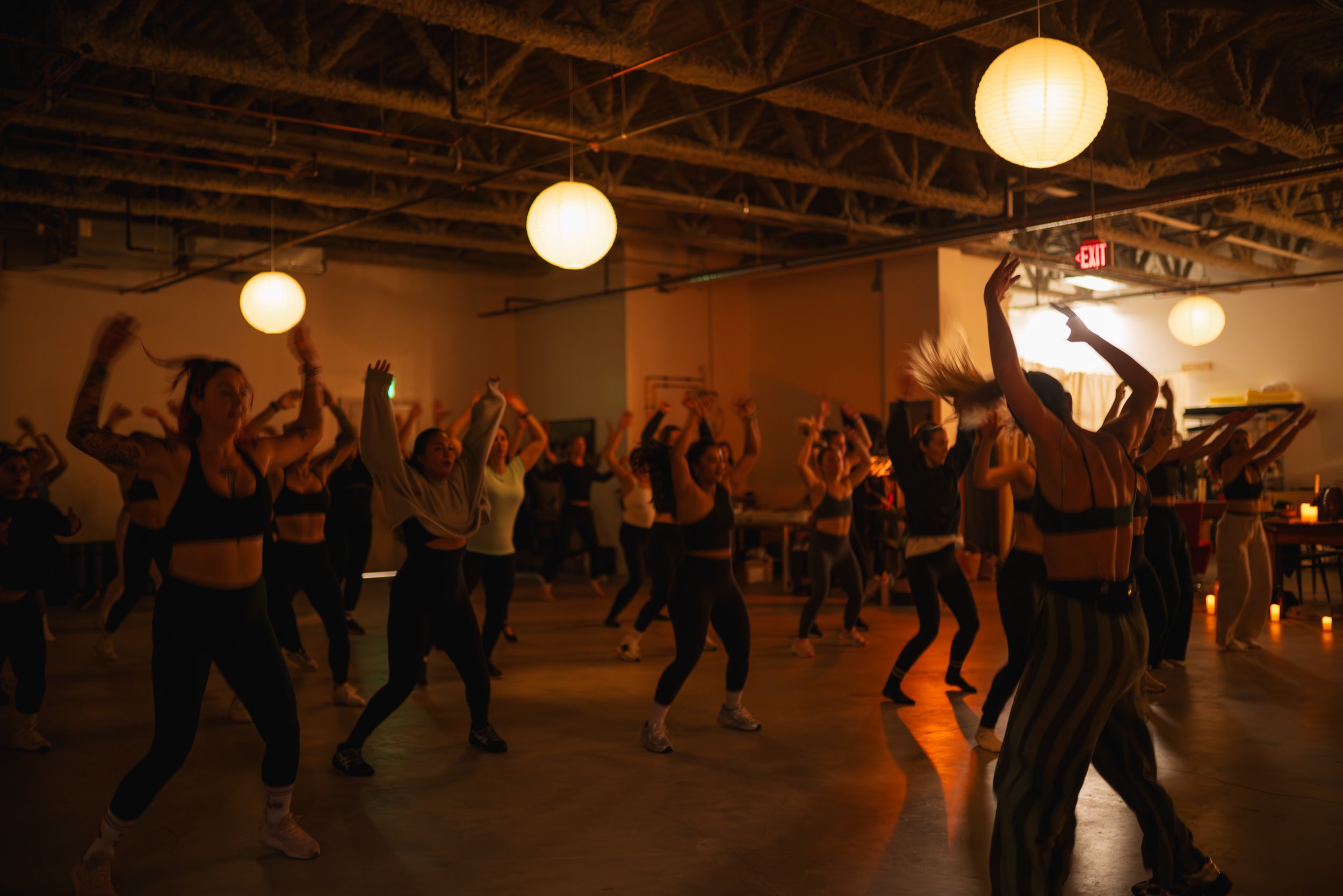 Group of people dancing in a dimly lit room with hanging paper lanterns, some with arms raised and hair moving.