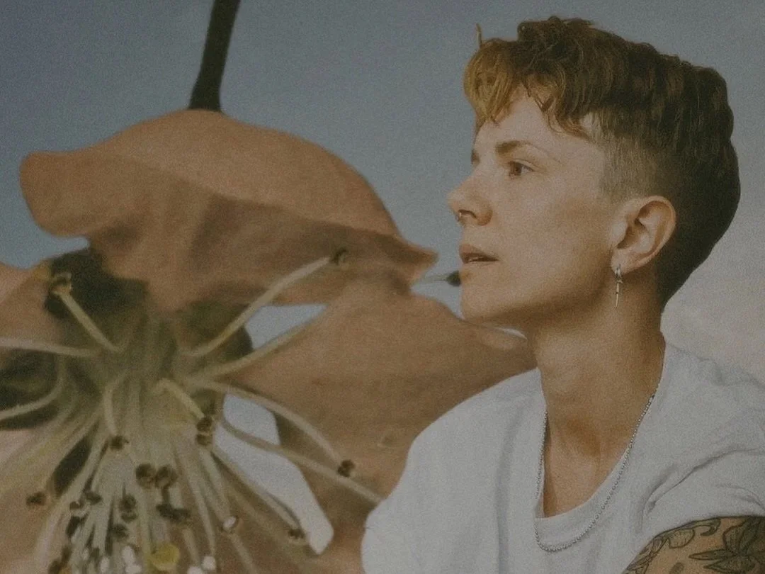 A young person with short, curly hair and earrings, wearing a white shirt and silver jewelry, looking at a large leafless plant or flower.