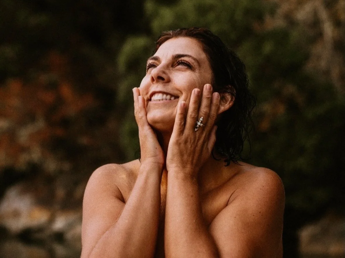 a woman with dark hair smiling and looking at the sky with her hands on her face. outside in nature, with wet hair