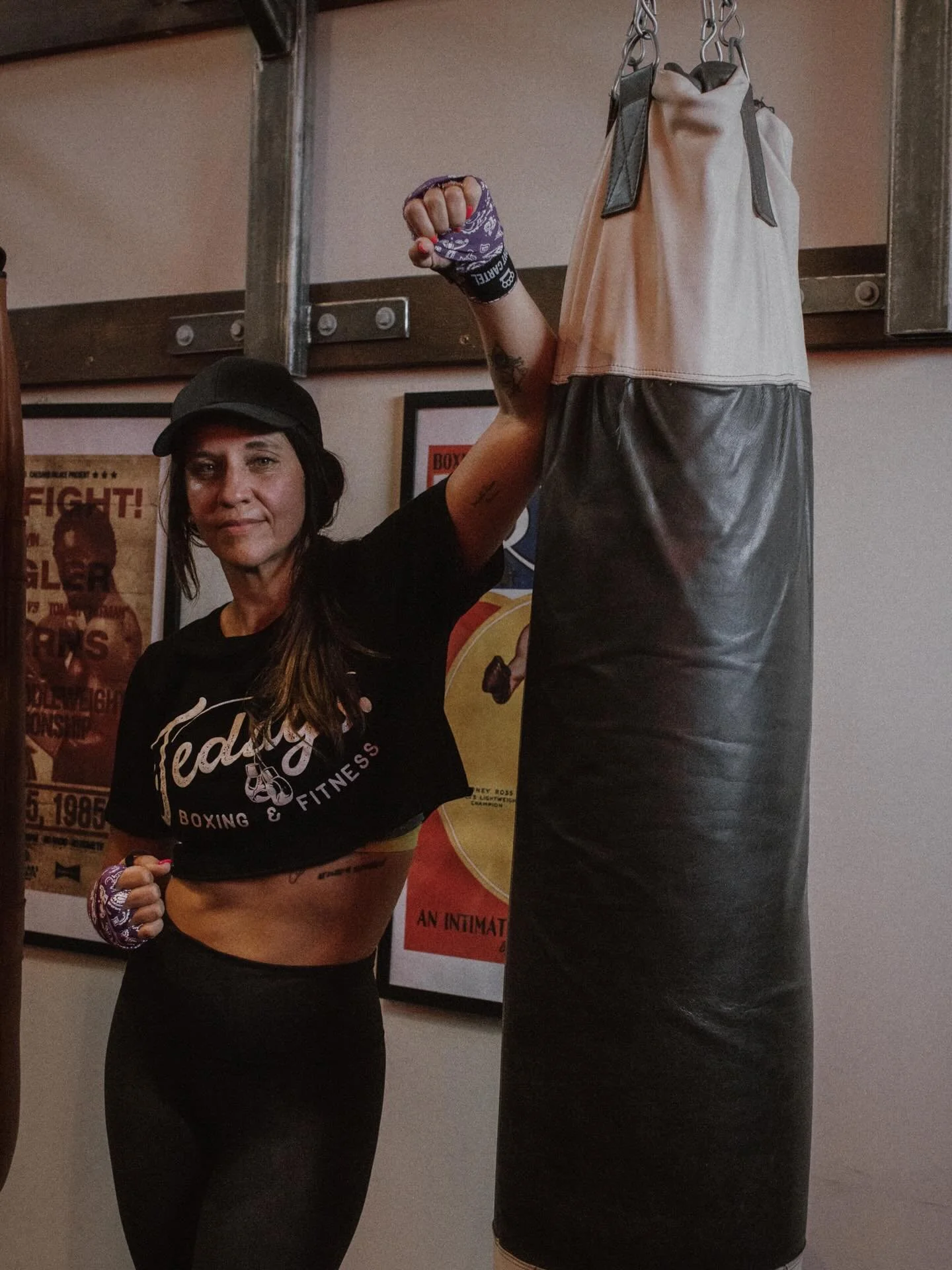 Female boxer in training stance with her arm raised next to a punching bag in a gym.