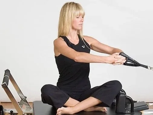 Woman sitting cross-legged on a pilates reformer, stretching her arms with resistance bands in a fitness studio.