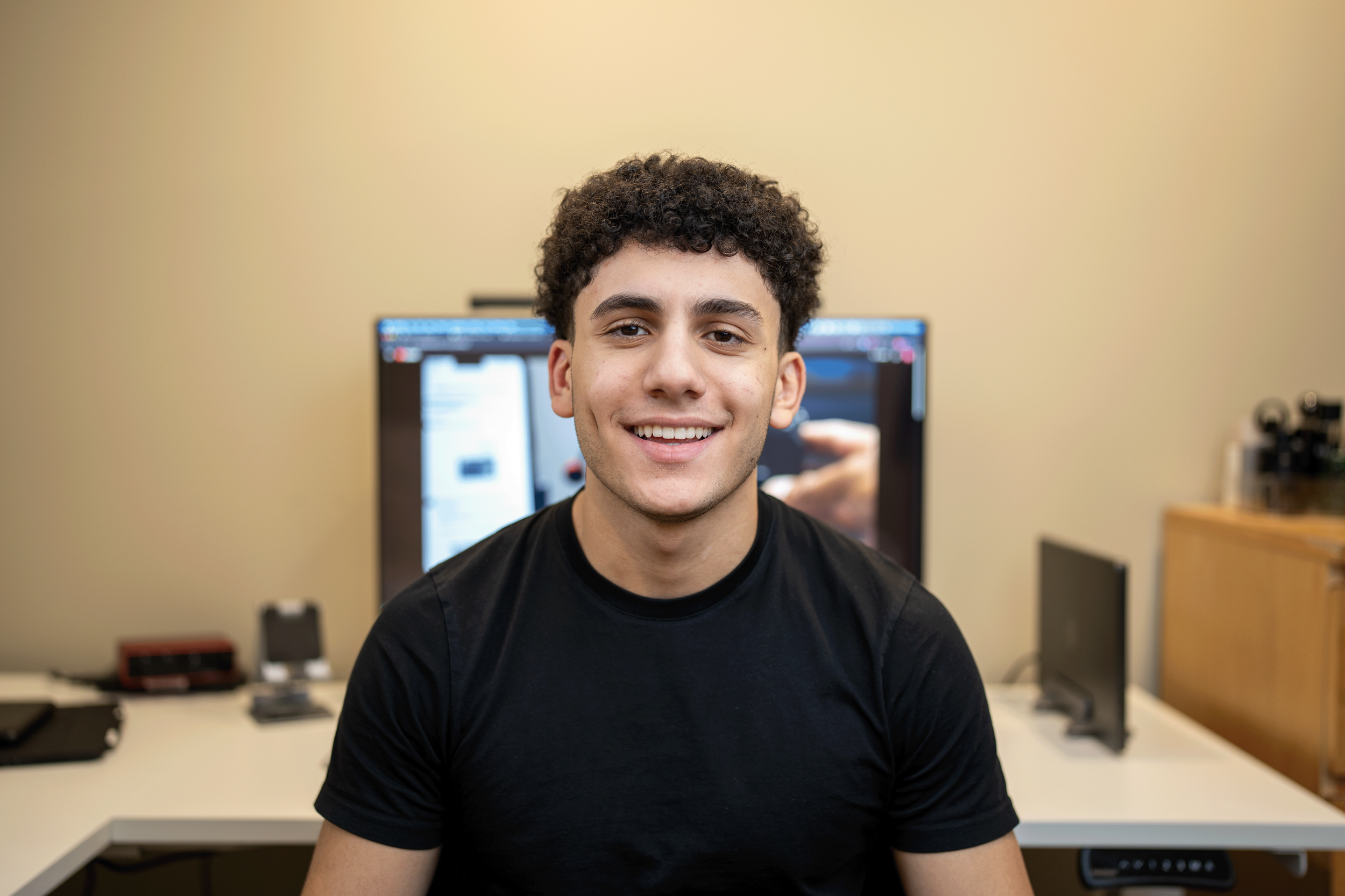 Young man sitting at desk with computer monitor behind him, smiling at camera.