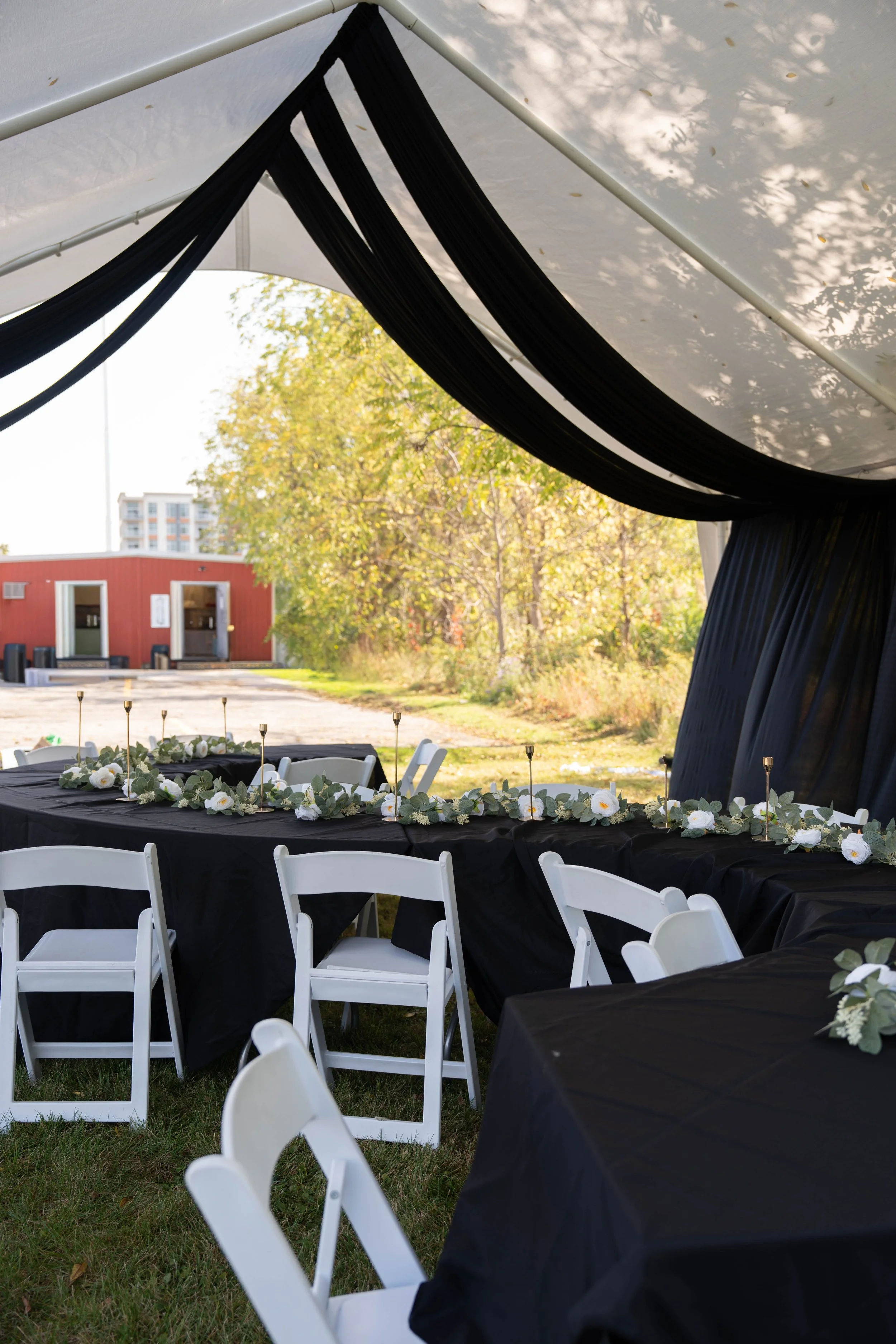 Outdoor event setup under a white tent with black drapery, featuring a long black table decorated with white flowers and green foliage, surrounded by white chairs, with trees and buildings visible in the background.