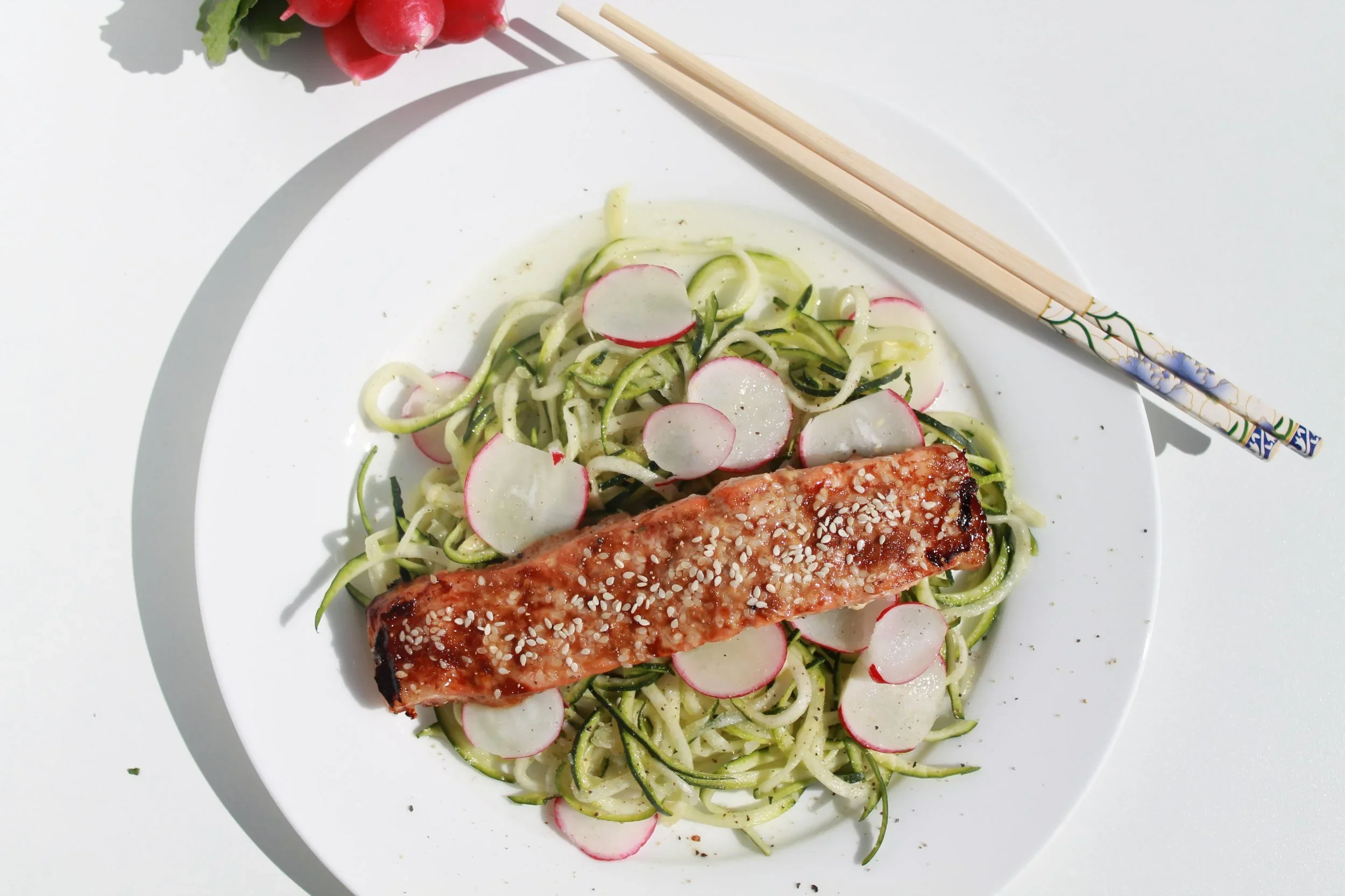 A white plate with spiralized zucchini and radish slices topped with a grilled salmon fillet sprinkled with sesame seeds, and a pair of decorative chopsticks resting on the plate.