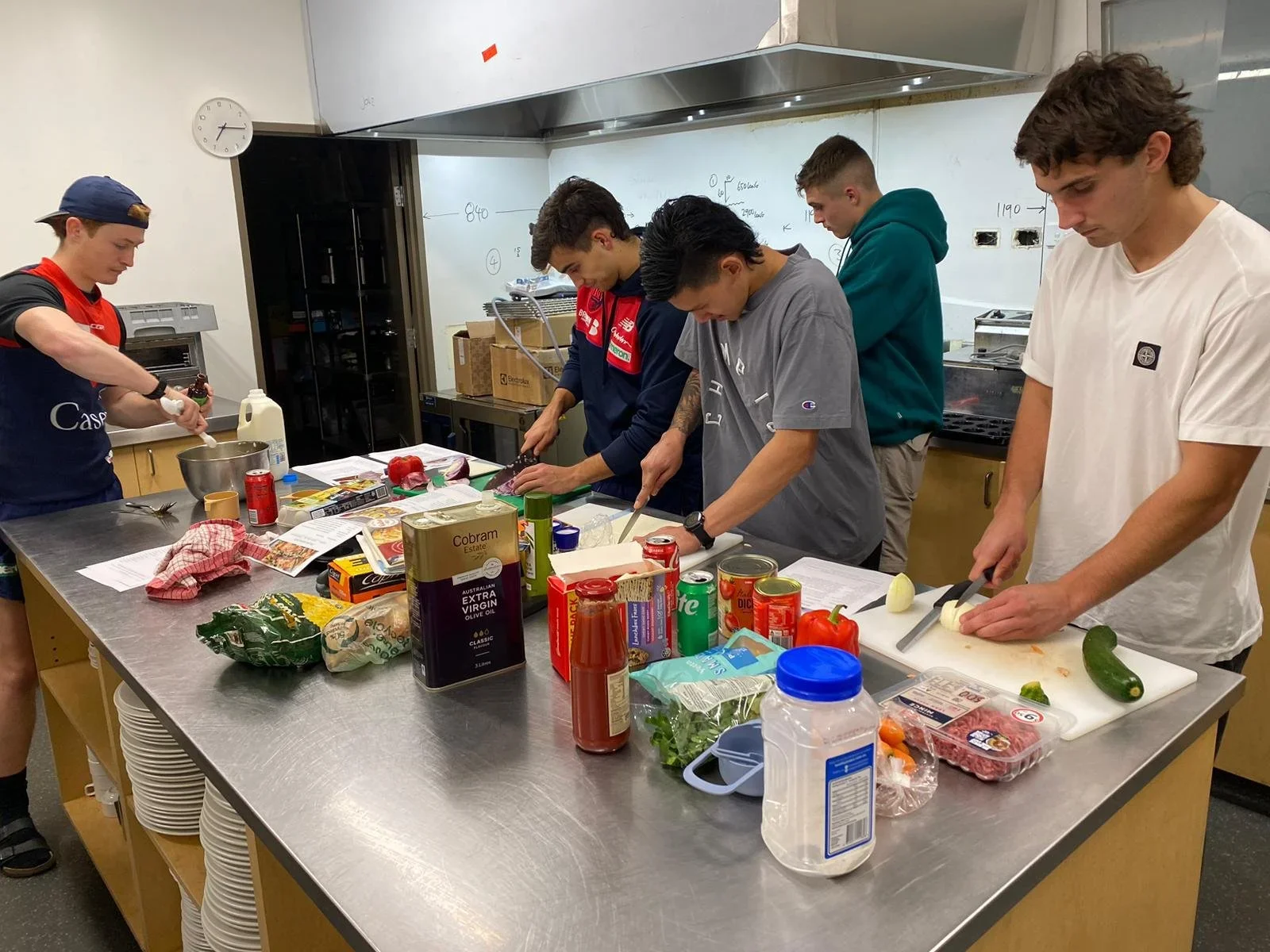 Five young men are cooking and preparing food together in a kitchen with a stainless steel countertop. They are chopping vegetables and handling various ingredients for a meal.