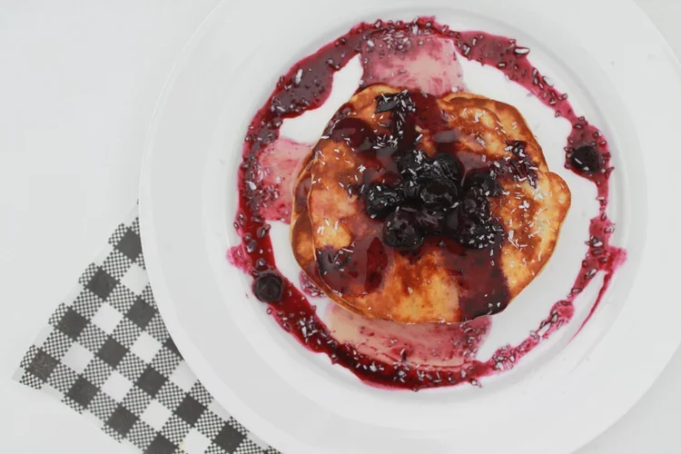 A dessert on a white plate with a pancake topped with blueberries and berry syrup, surrounded by more berry syrup and crumbs, on a checkered napkin.