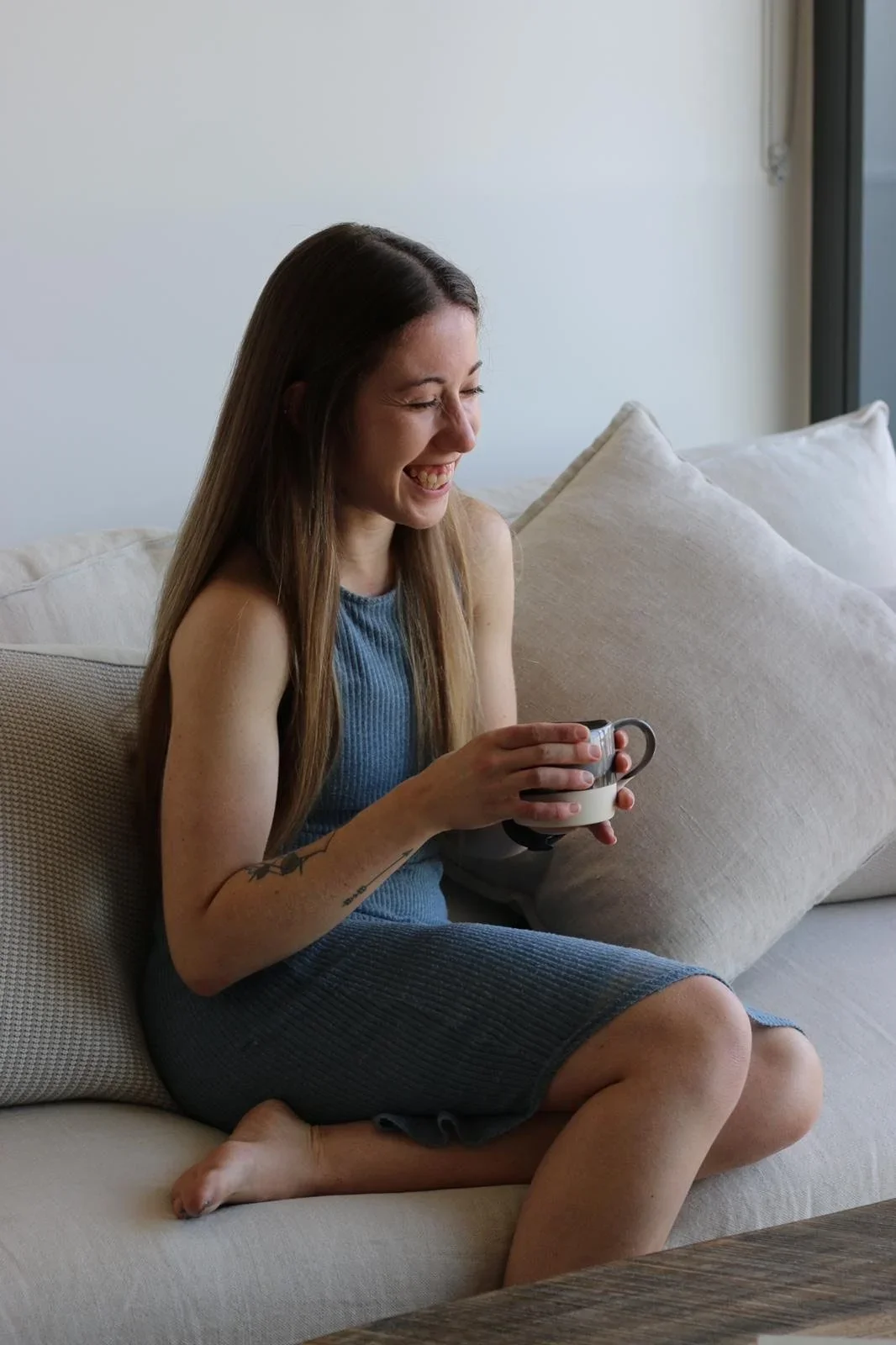 A woman with long brown hair and tattoos on her arm, wearing a blue sleeveless dress, sitting cross-legged on a beige couch, holding a coffee mug, and laughing.