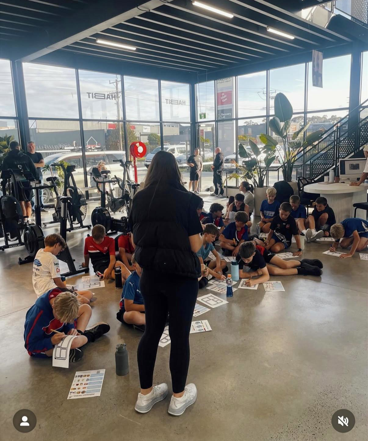 Group of children sitting on the floor of a gymnasium, doing worksheets, with an instructor standing nearby, in a fitness center with exercise bikes and large windows.