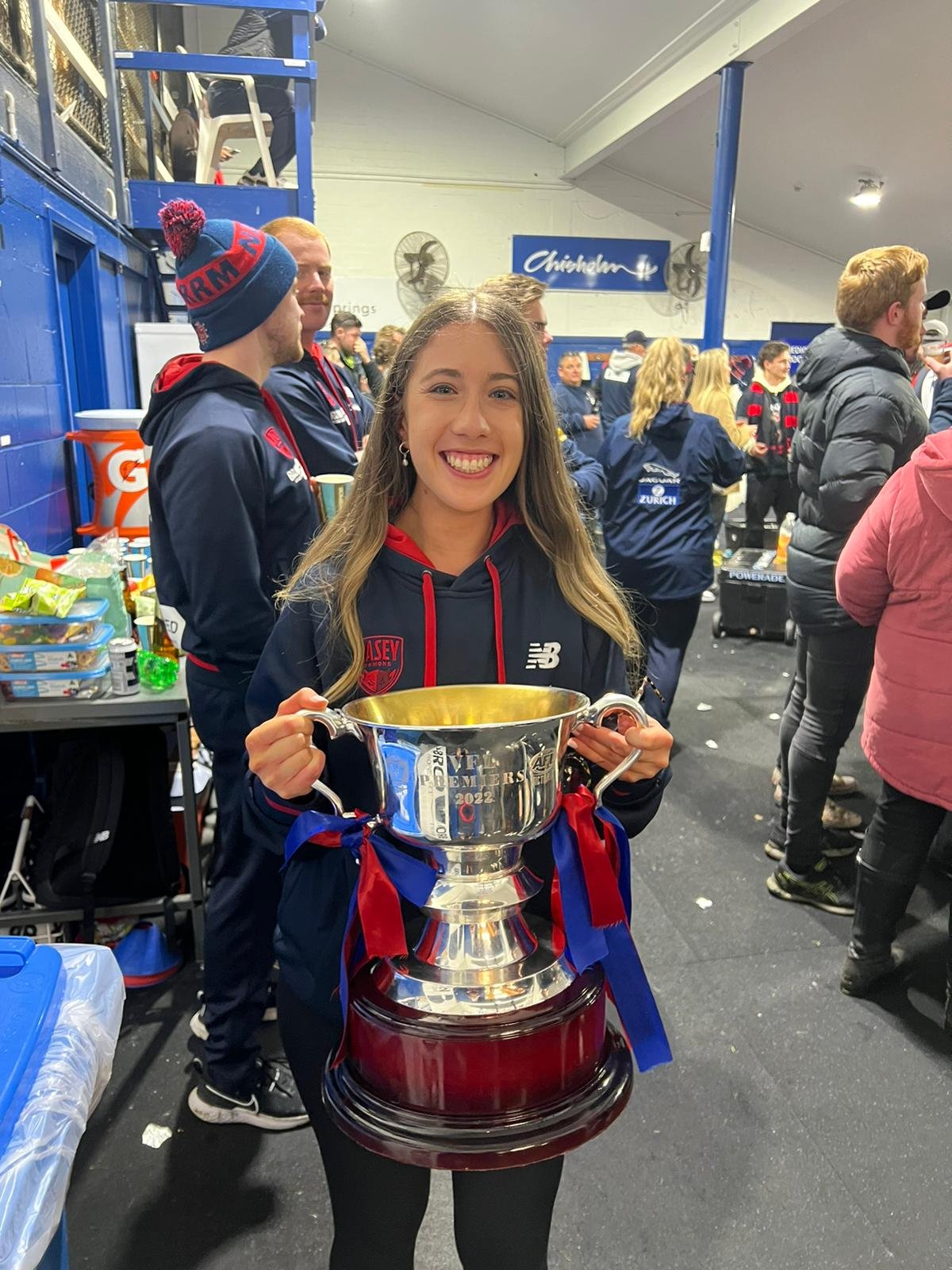 A young woman holding a large silver trophy with red and blue ribbons, smiling at the camera in a crowded sports facility.