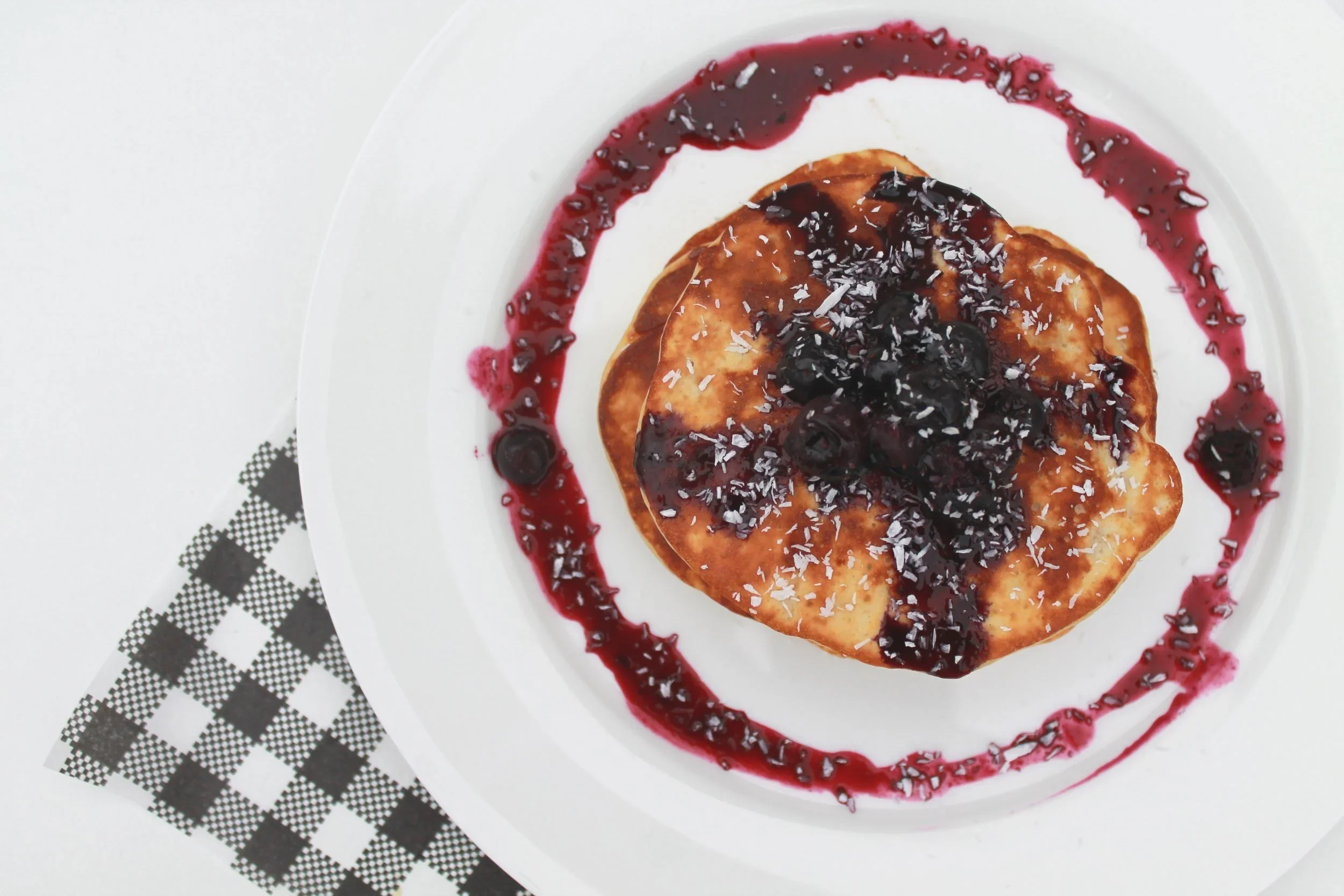 Pancakes topped with blueberry compote and drizzled with berry sauce on a white plate.