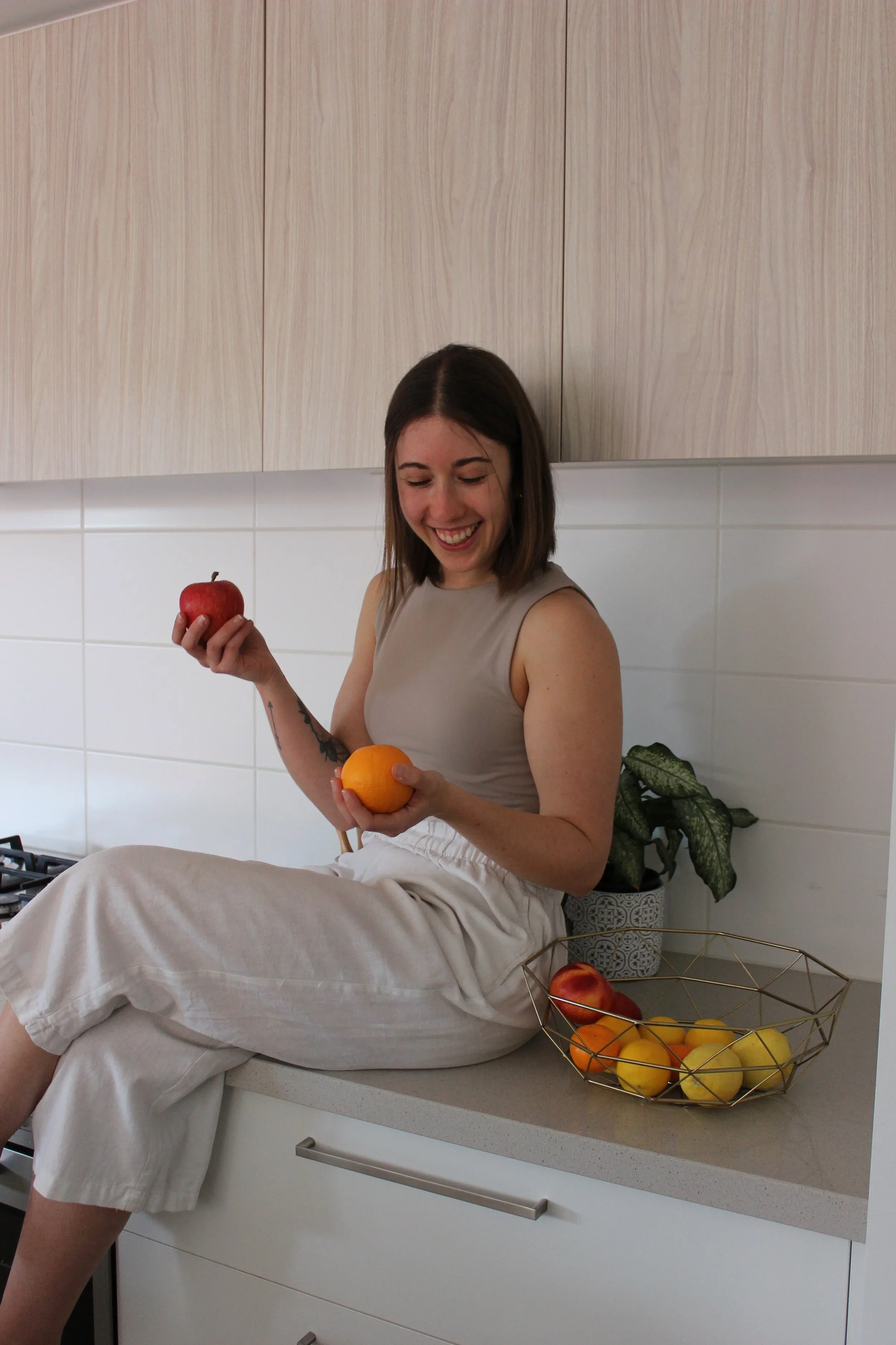 A woman sitting on a kitchen countertop holding an apple in her right hand and an orange in her left hand, smiling down at the fruits. There is a wire basket with yellow and red fruits and a potted plant on the counter beside her.