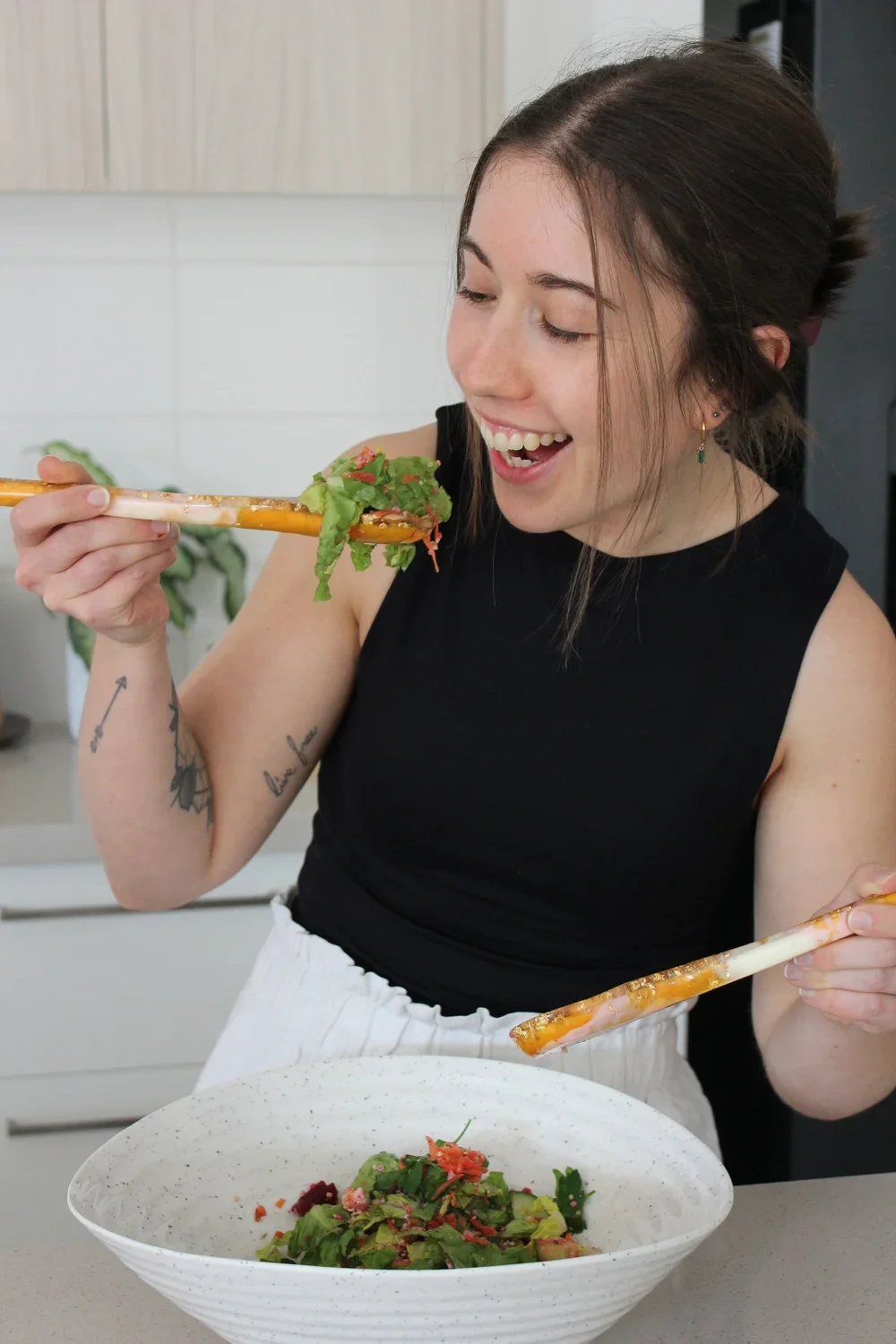 A woman with brown hair, earrings, and tattoos on her arms is smiling while using chopsticks to pick up a salad from a white bowl, in a kitchen setting.