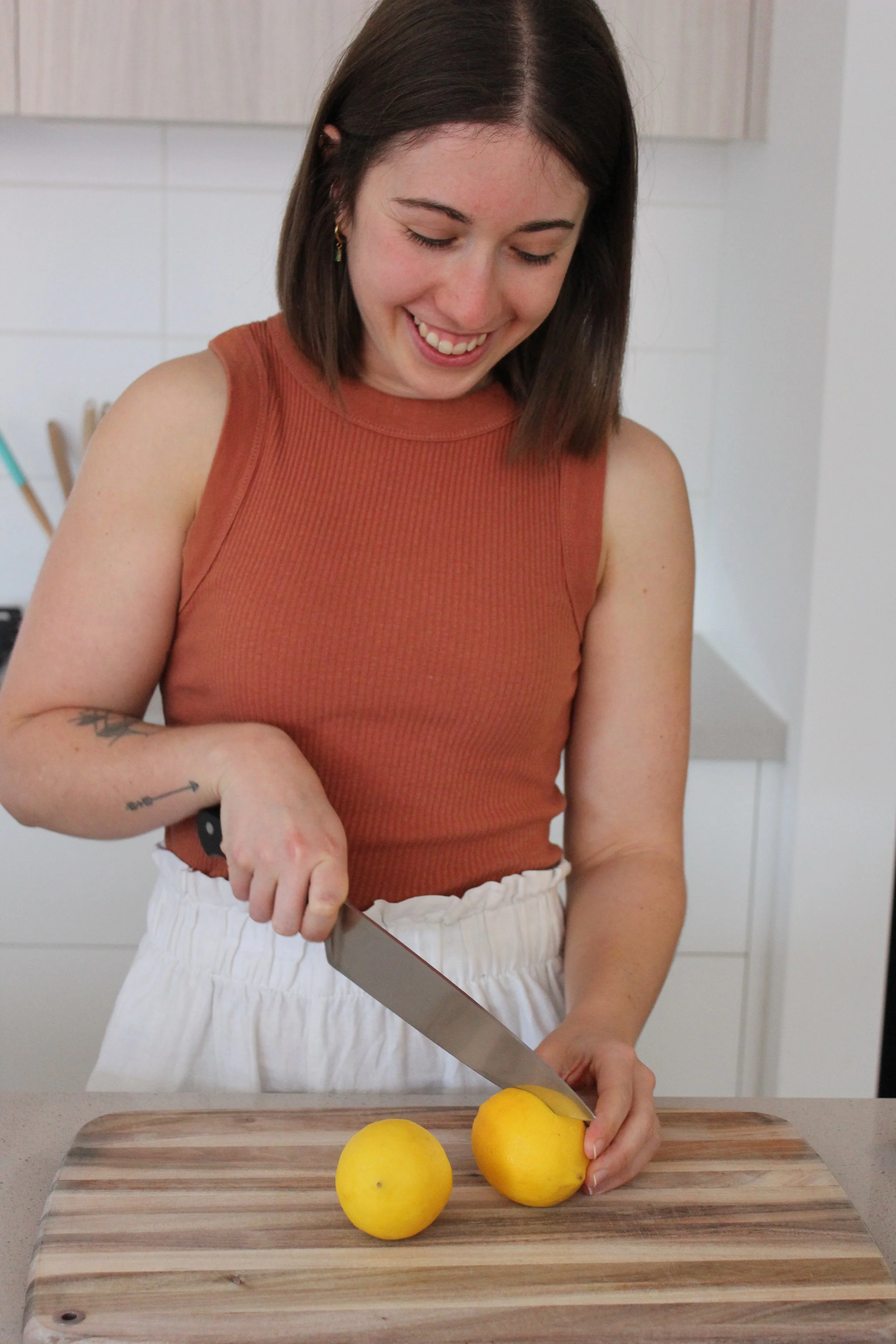A woman with shoulder-length brown hair, wearing a sleeveless orange top, is smiling as she slices a lemon on a wooden cutting board in a kitchen.
