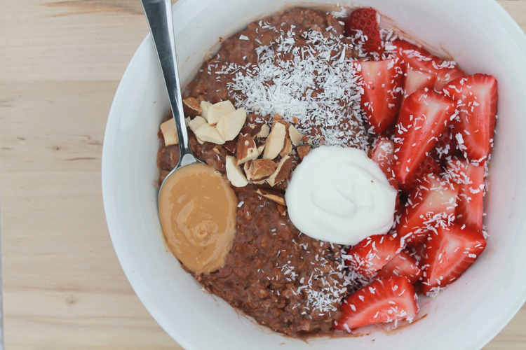 Chocolate oatmeal topped with sliced strawberries, shredded coconut, chopped nuts, a dollop of whipped cream, and a spoonful of peanut butter in a white bowl on a wooden surface.